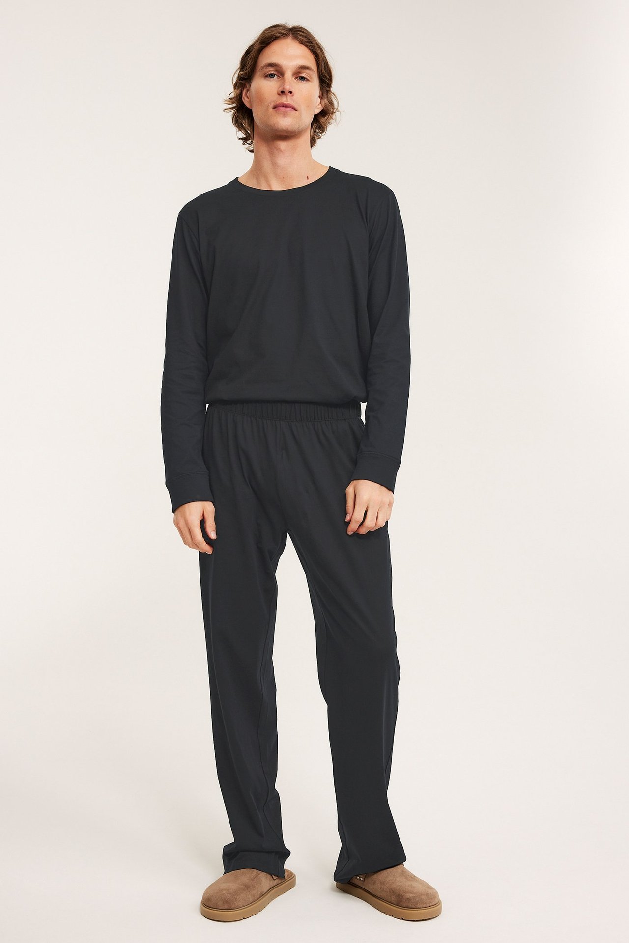 Framifrån: man i svarta bomullsjersey pyjamasbyxor med resår, svart långärmad tröja och bruna tofflor.