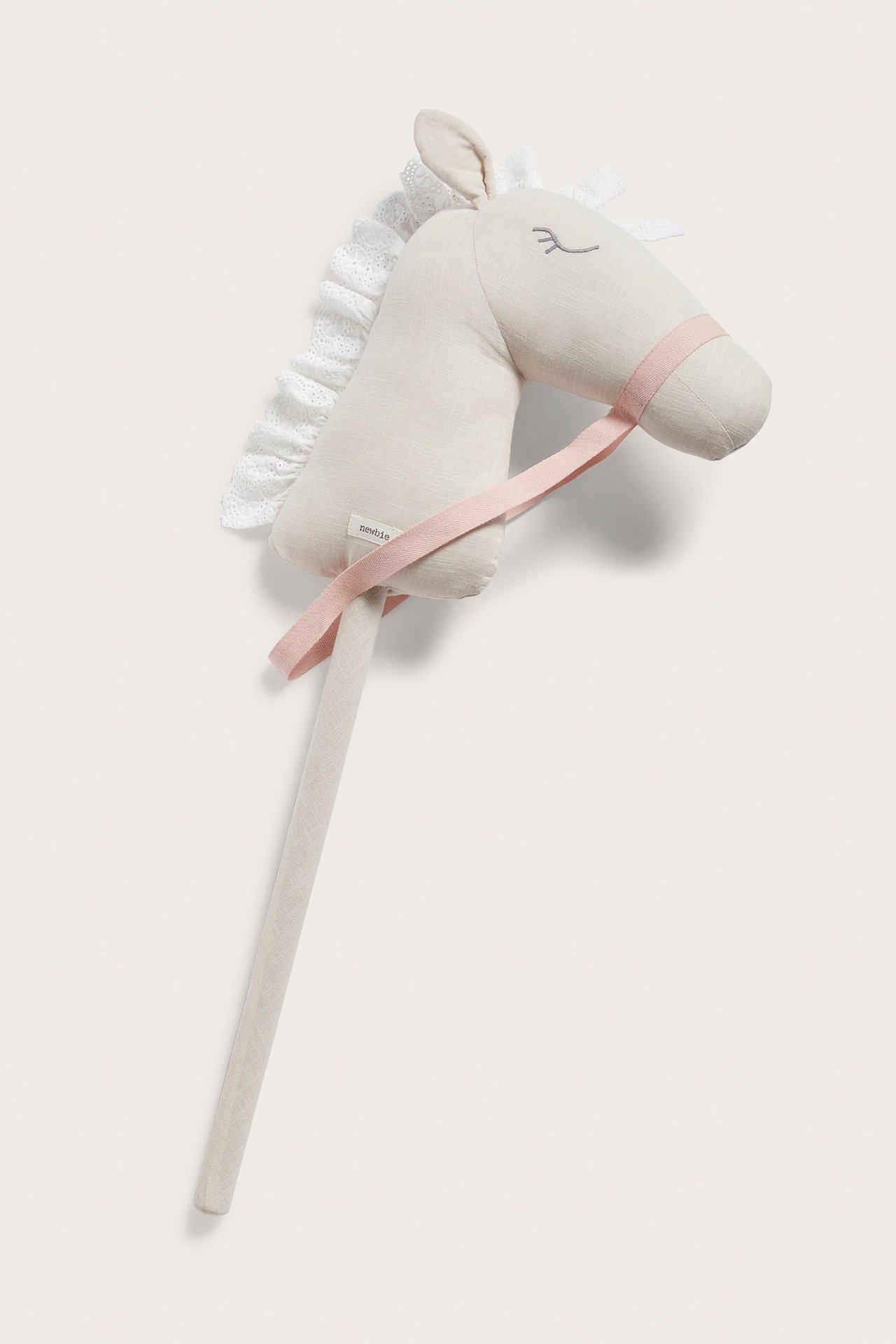 Widok z przodu: beżowy konik na patyku z bawełny organicznej, biała koronkowa grzywa, haftowane oczy, różowa uzda.