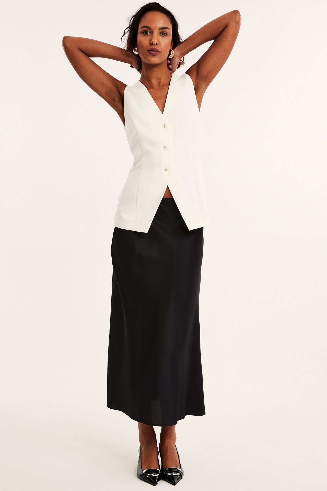 Widok z przodu: Kobieta w czarnej rozkloszowanej satynowej spódnicy midi, białej kamizelce i czarnych szpilkach.