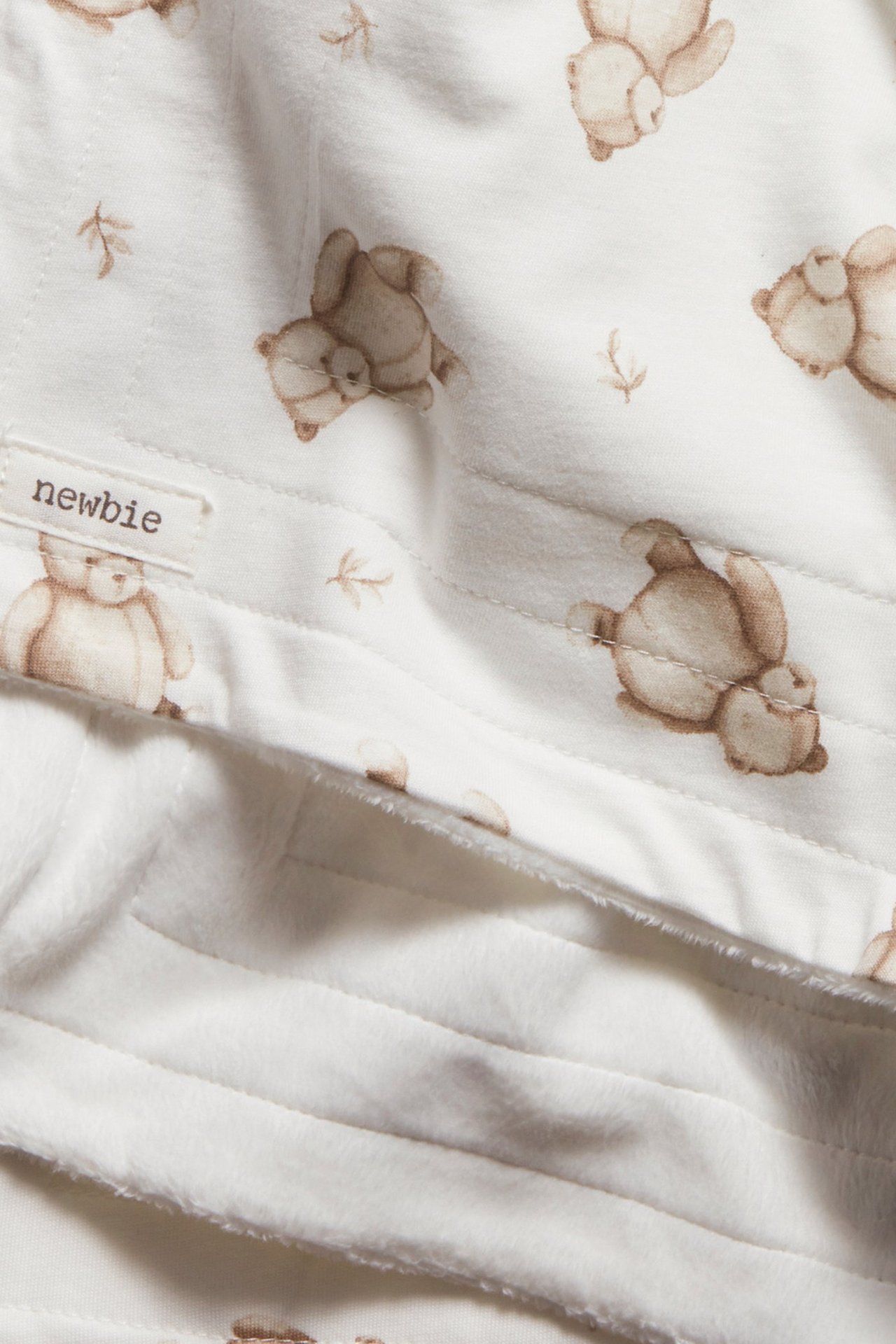 Nærbilde av et offwhite babyteppe med brunt teddybjørnmønster og myk fleeceside, med Newbie-etikett.