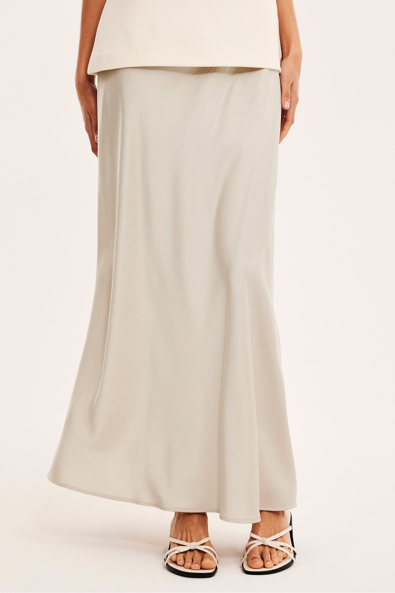 Frontbilde av kvinne i et lys beige utsvingt satengskjørt, en kremfarget topp og hvite sandaler.