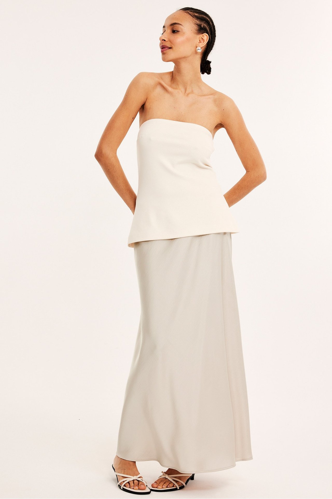 Widok z przodu kobiety w długiej, rozkloszowanej jasnobeżowej satynowej spódnicy, kremowym topie i jasnych sandałach.