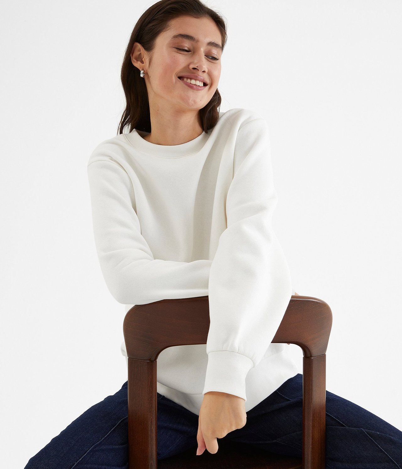 Widok z przodu kobiety w kremowej bluzie z okrągłym dekoltem, ciemnoniebieskich dżinsach i małych srebrnych kolczykach.