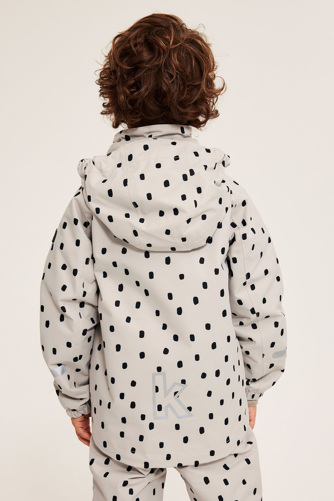 Barn i beige vanntett skalljakke med svarte prikker, sett bakfra.