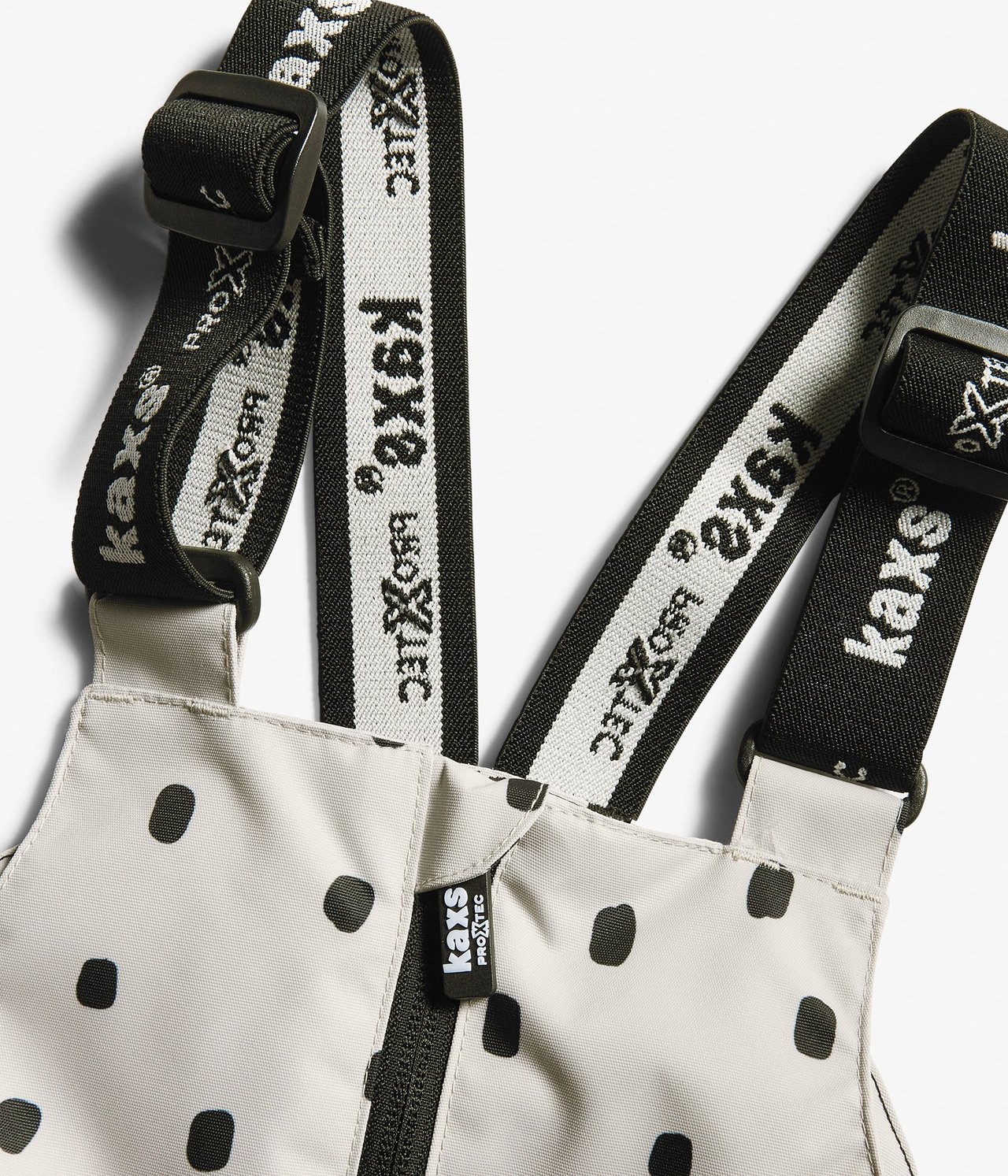 Zbliżenie na beżowe, w kropki spodnie dziecięce z czarno-białymi szelkami z logo i zamkiem błyskawicznym.