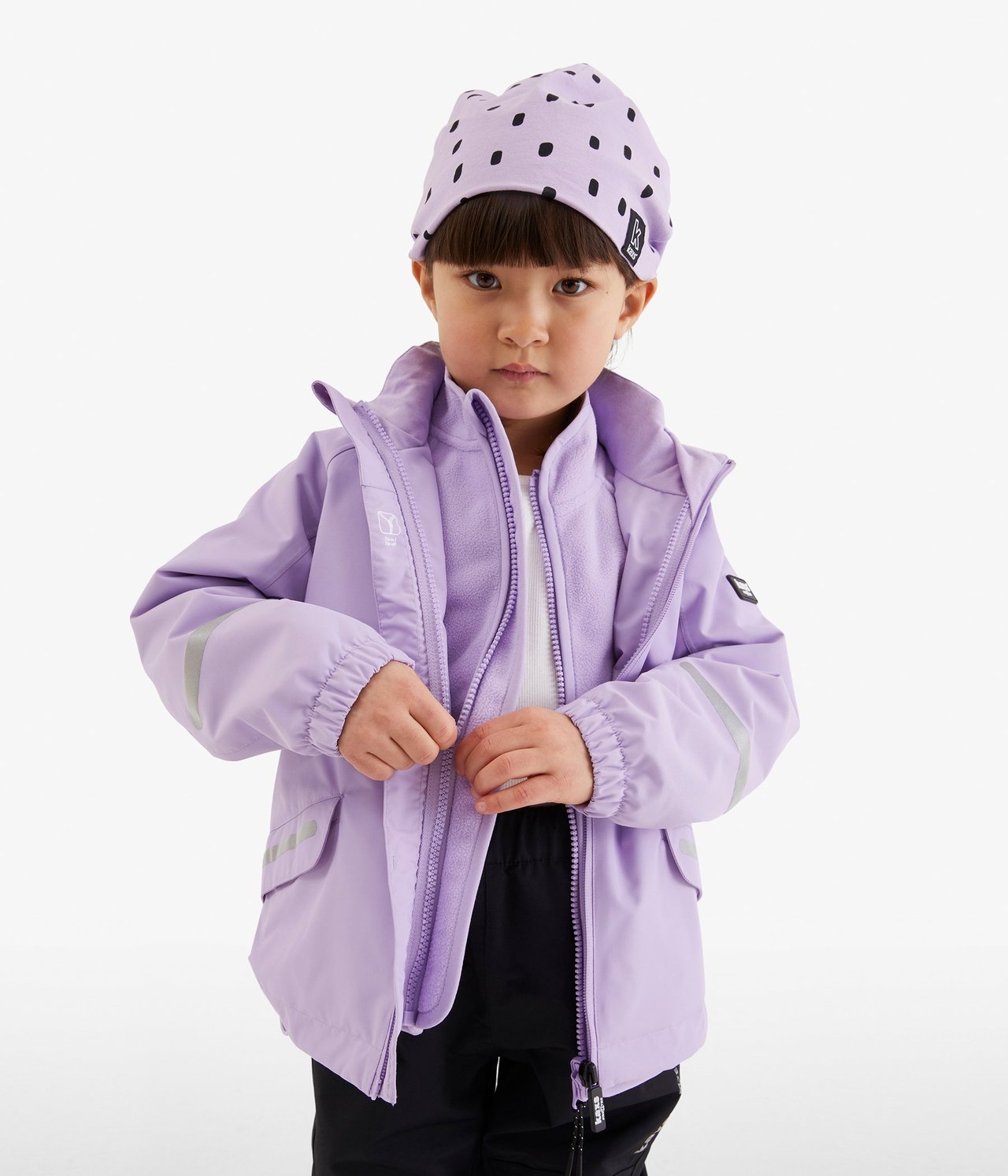 Widok z przodu: Dziecko zapina liliową kurtkę shellową na polar, z czarnymi spodniami i liliową czapką w kropki.