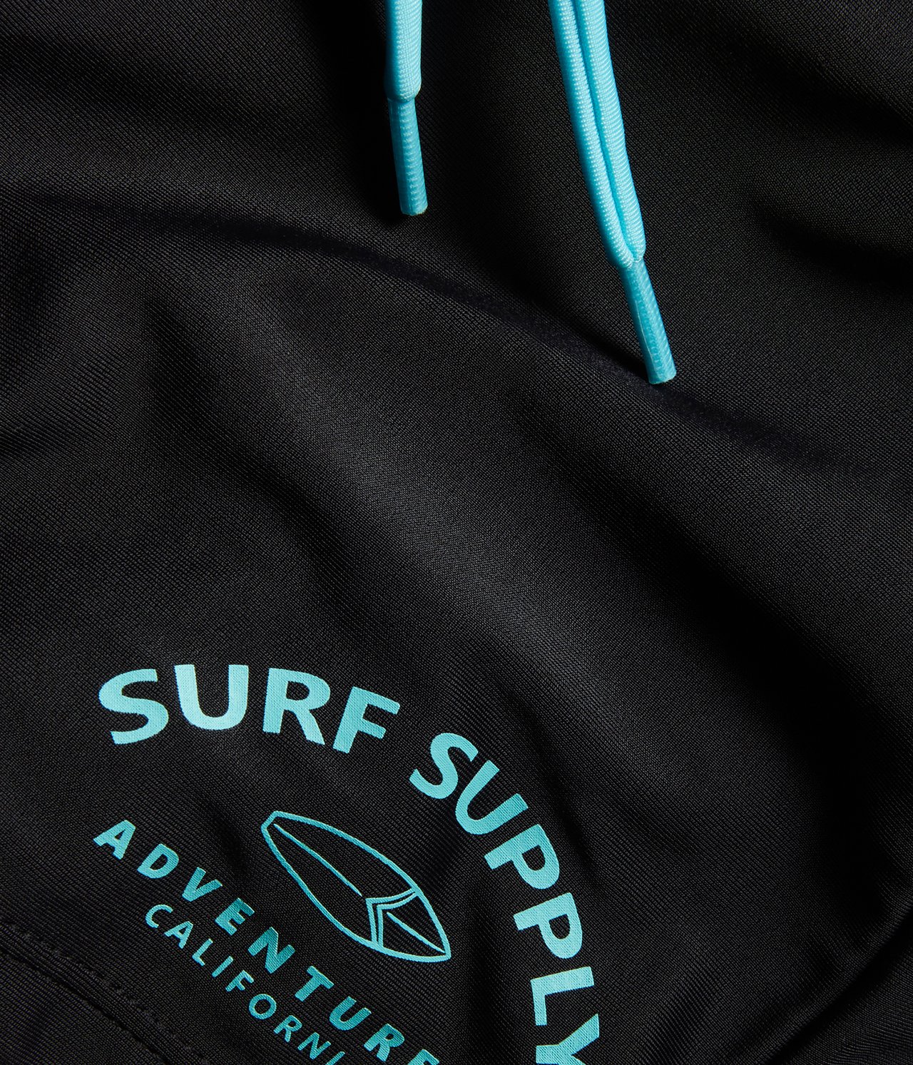 Närbild av svarta badbyxor med ljusblå dragsko och ett "SURF SUPPLY"-tryck med surfbräda.