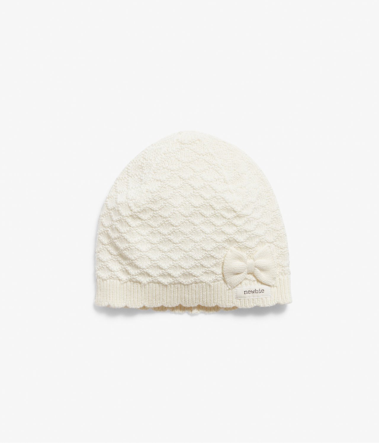 Framsida av en offwhite mönsterstickad baby mössa med liten rosett och ribbad vågig kant.