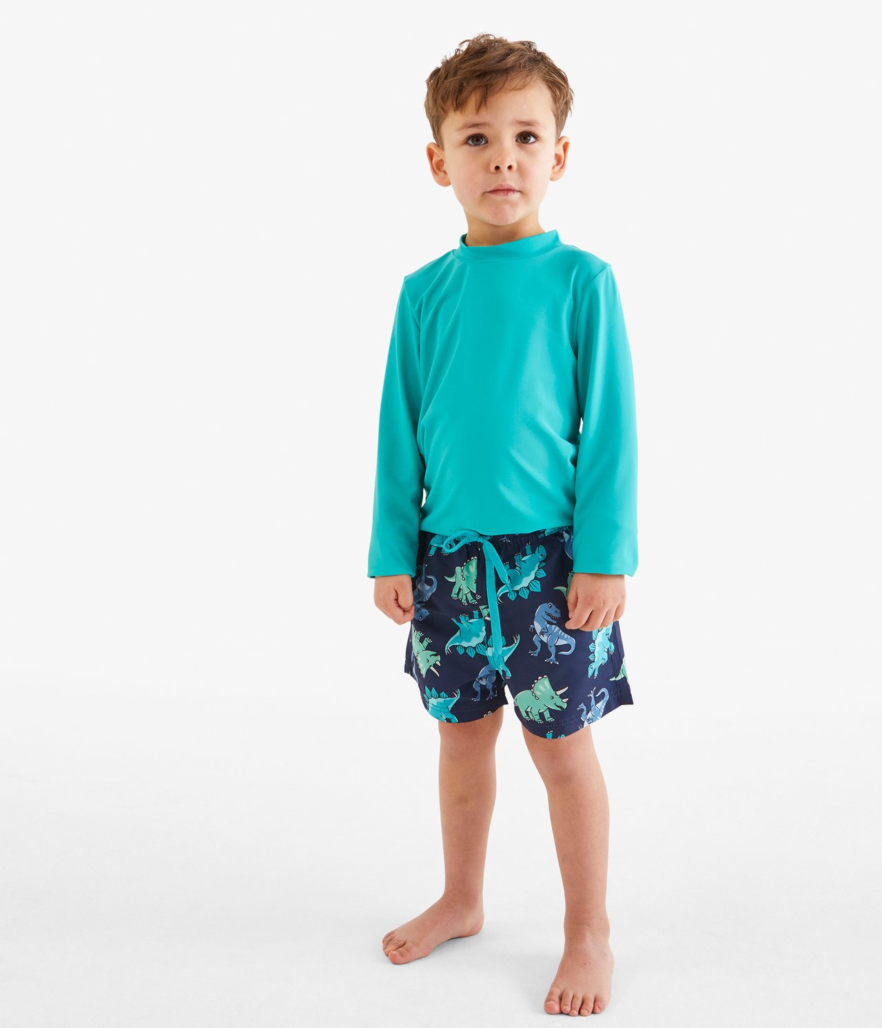 Framifrån: Ett barn i turkos långärmad UV-tröja och mörkblå badshorts med dinosauriemönster.