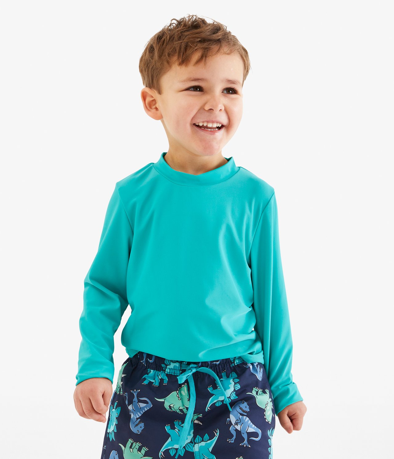 Framifrån, leende barn i turkos långärmad UV-tröja och mörkblå badshorts med dinosauriemönster.