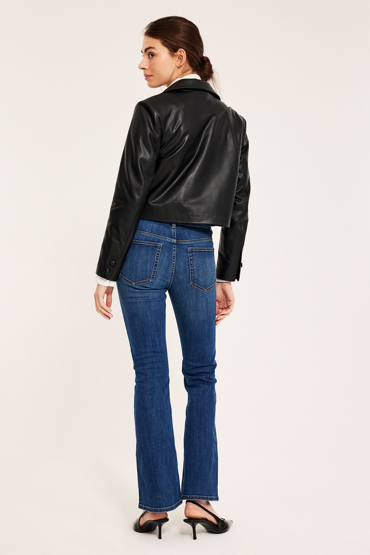 Naisen takaa-sivukuva: denim flare-farkut, musta nahkatakki valkoisen paidan päällä, mustat korot.