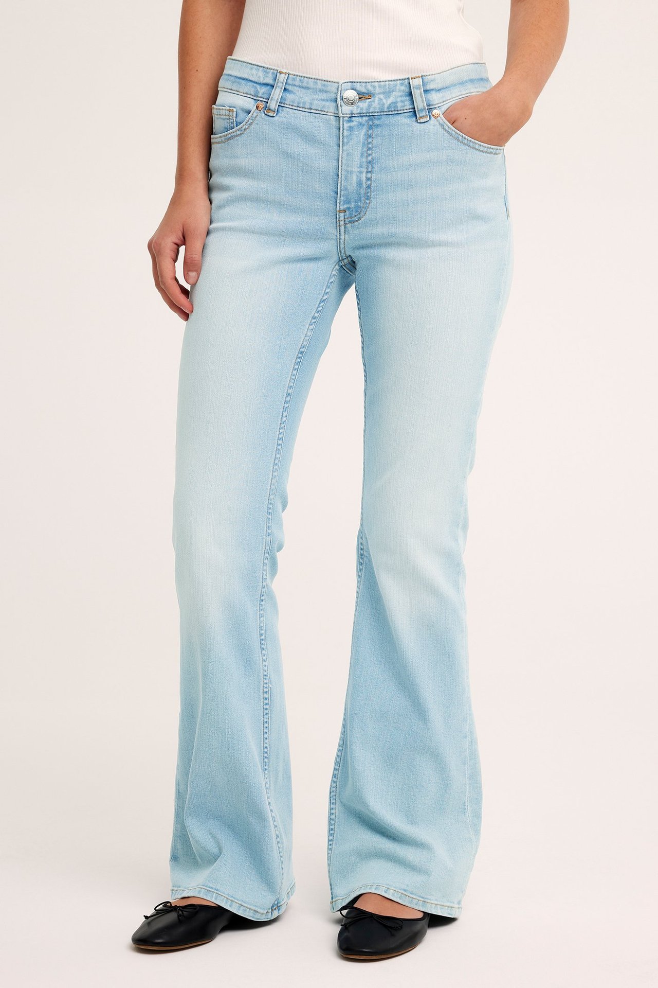 Frontvy av en kvinna i ljusblå lågmidjade utsvängda jeans, ett vitt ribbat linne och svarta ballerinaskor.