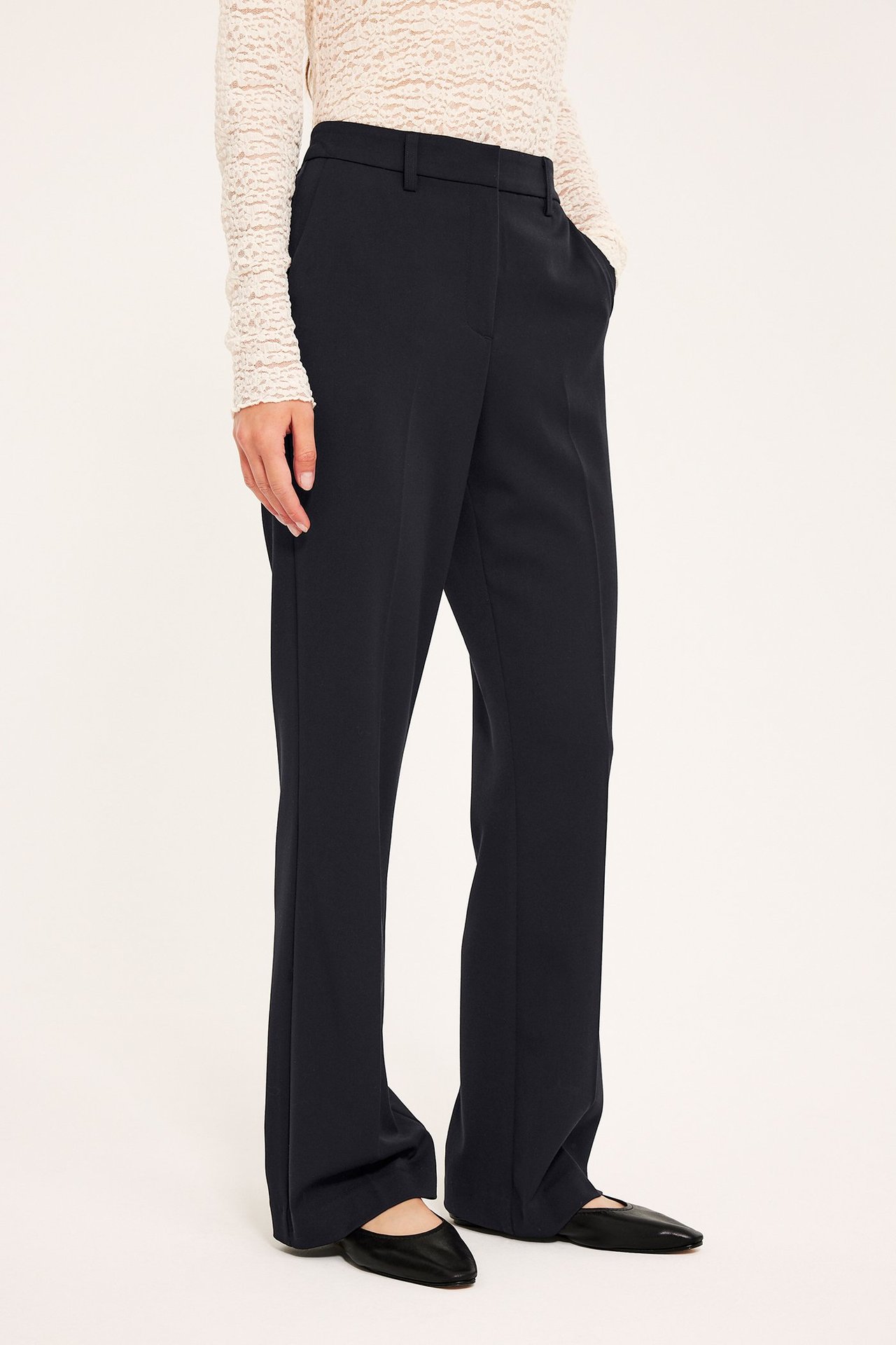 Forfra, kvinne i svarte smale utsvingte dressbukser, hvit blondetopp og svarte flate sko.