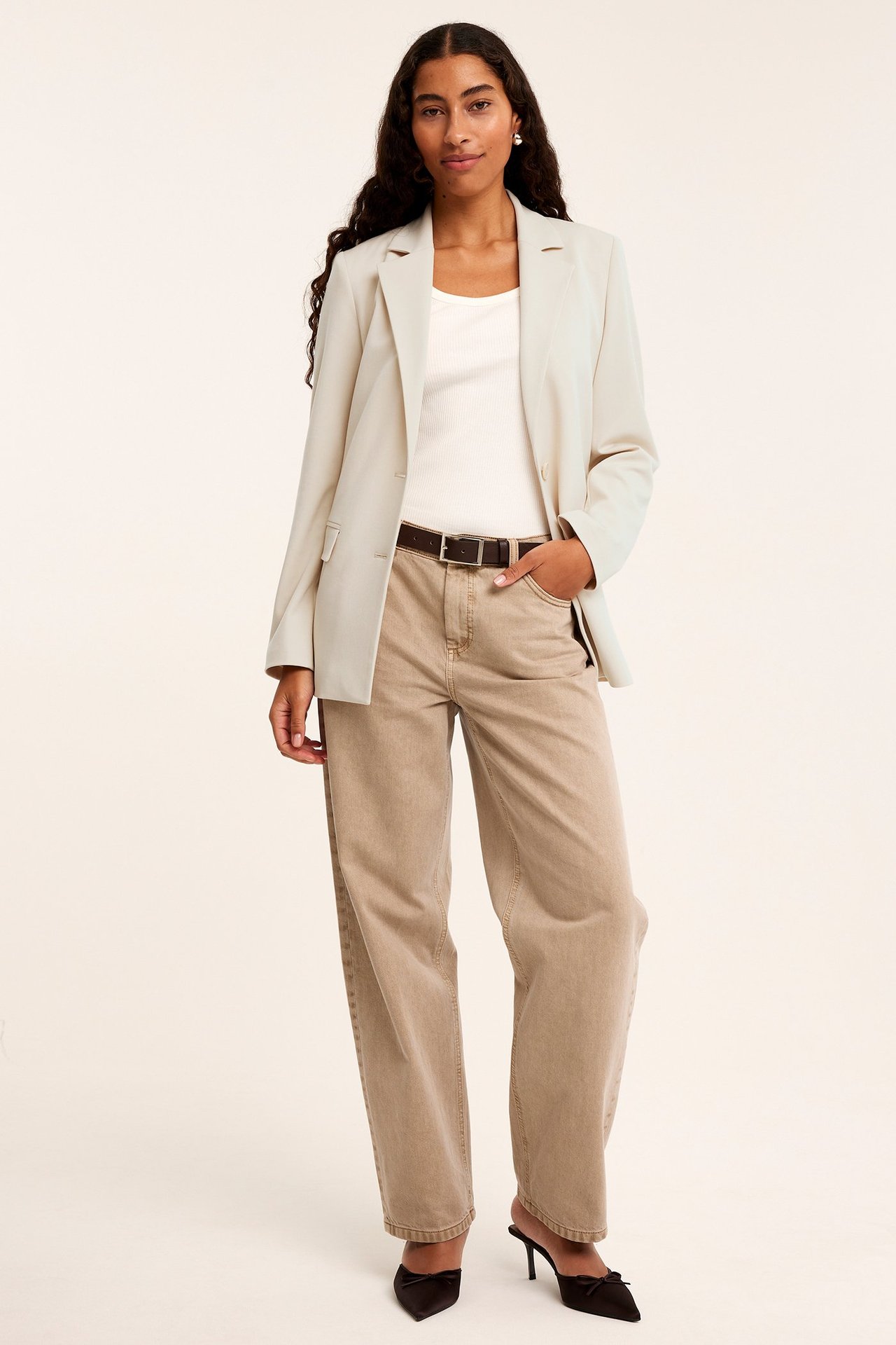 Edestä: nainen vaaleassa beigessä bleiserissä, valkoisessa topissa, beigessä leveässä housussa, ruskeassa vyössä, mustissa muuleissa.