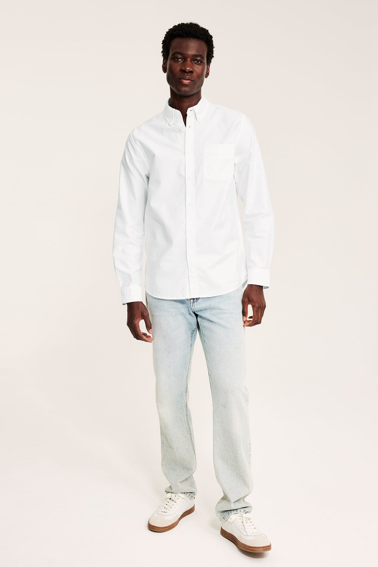 Fullfigur forfra av en mannlig modell iført en hvit langermet skjorte, lyse jeans og joggesko.
