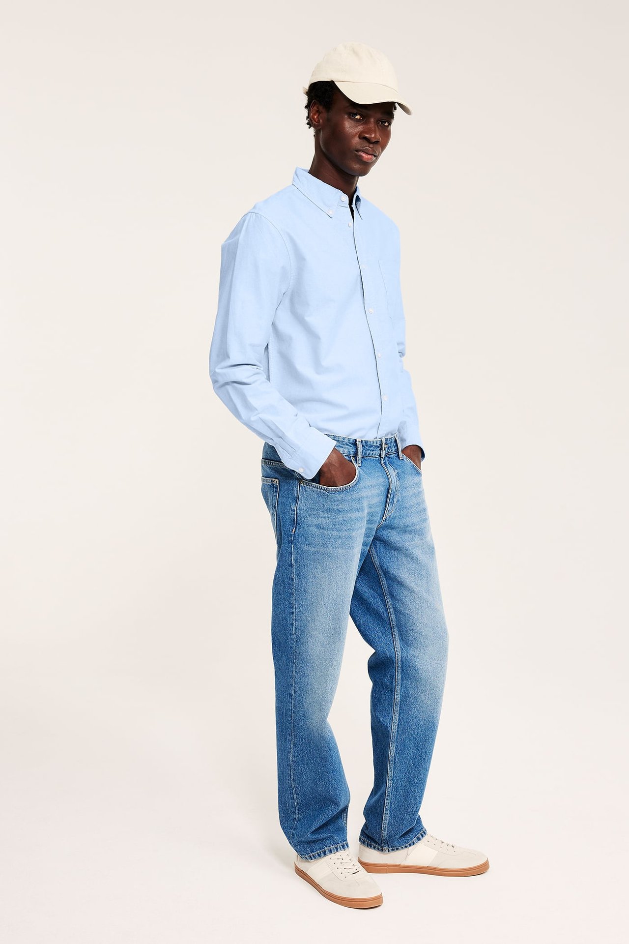 Frontvy av en man i en ljusblå oxfordskjorta, blåa jeans, beiga sneakers och en beige keps.