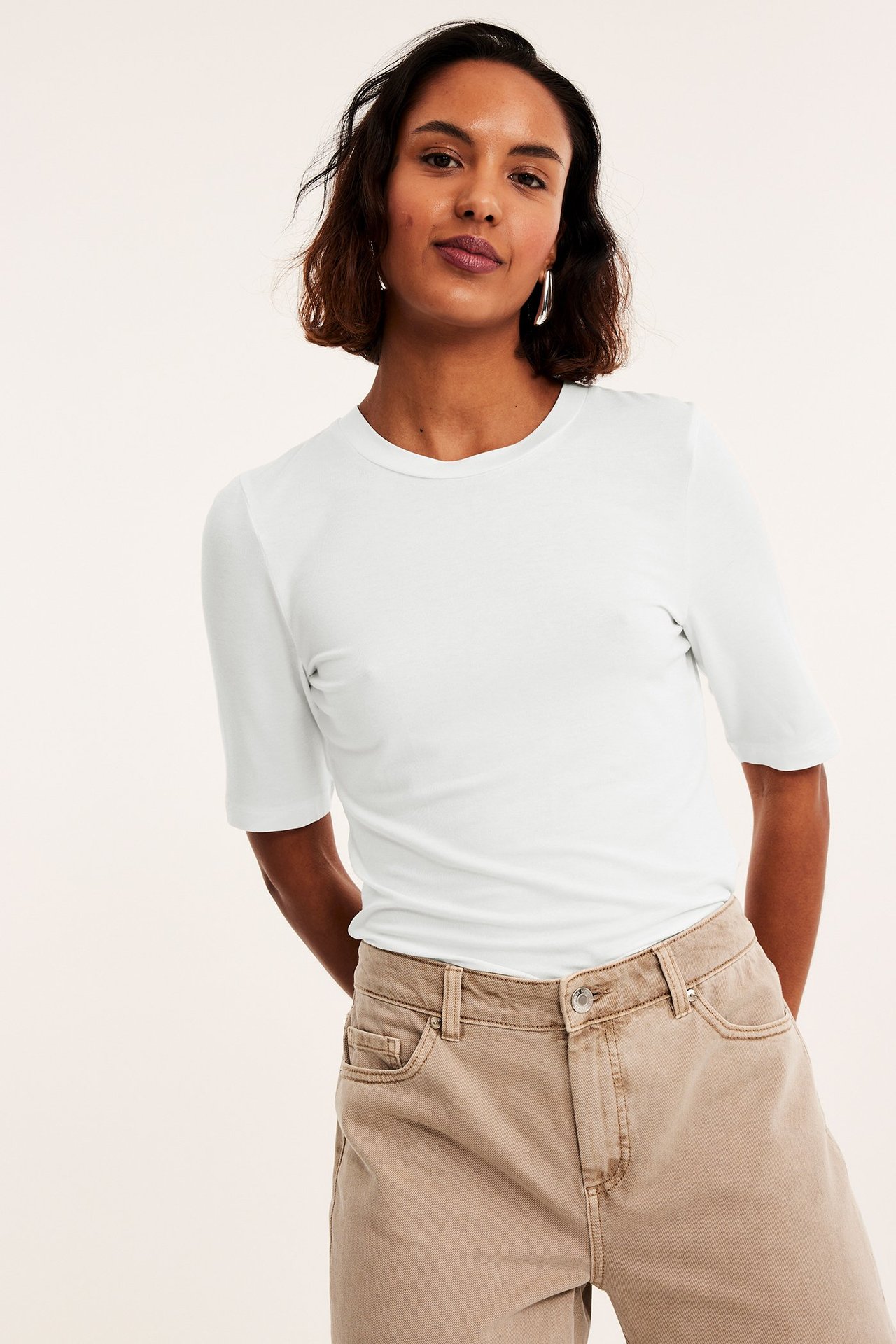 Forfra: Kvinne i en off-white, tettsittende viskose-t-skjorte med albuelange ermer og brune, vide bukser.