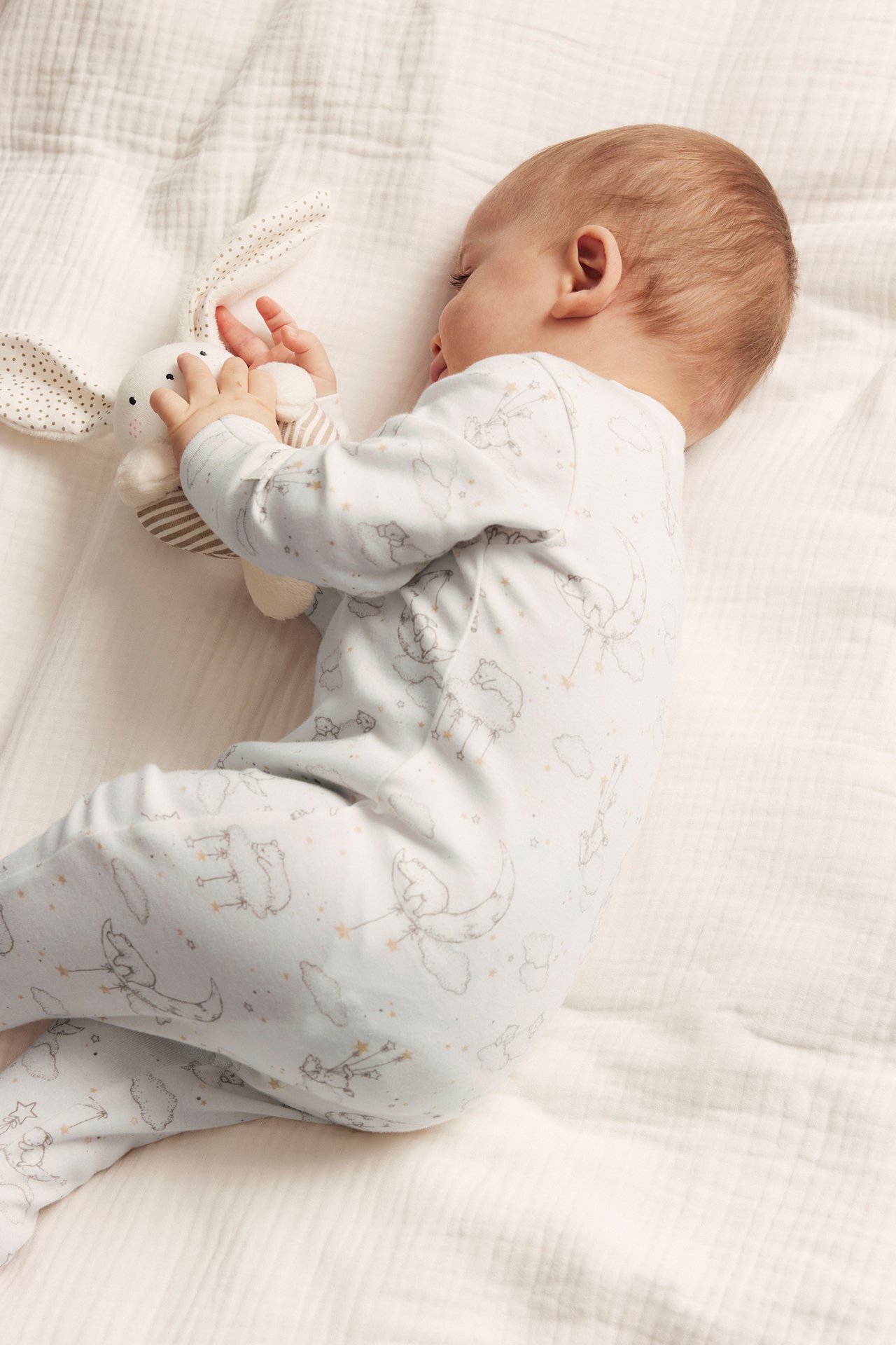 Sivukuva vauvasta offwhite-puuvillapyjamassa, jossa karhu- ja kuukuvio, pitäen valkoista kangaskania.