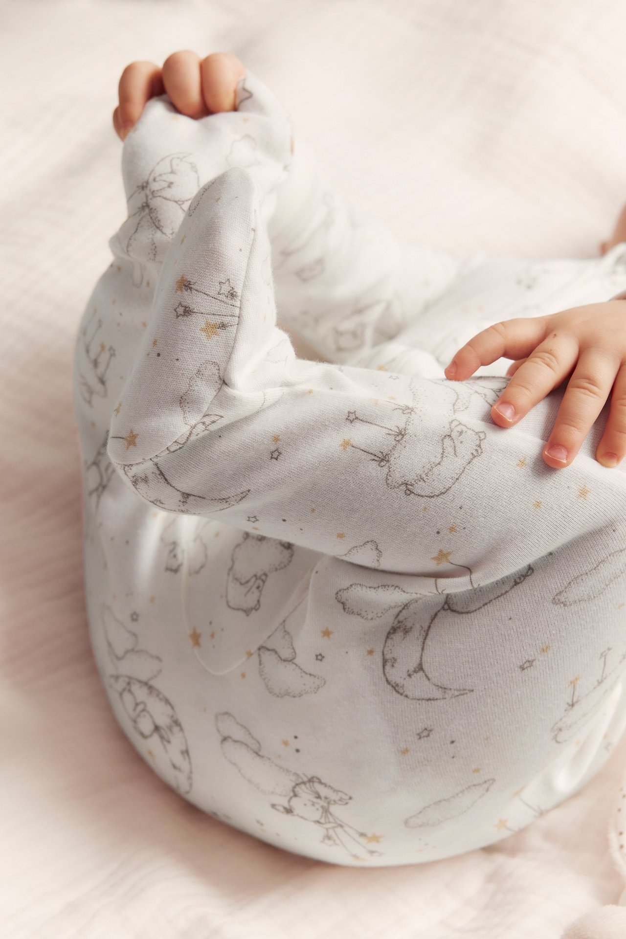 Närbild på baby i offwhite bomullspyjamas med grått djur-, mån-, stjärnmönster och integrerade fötter.
