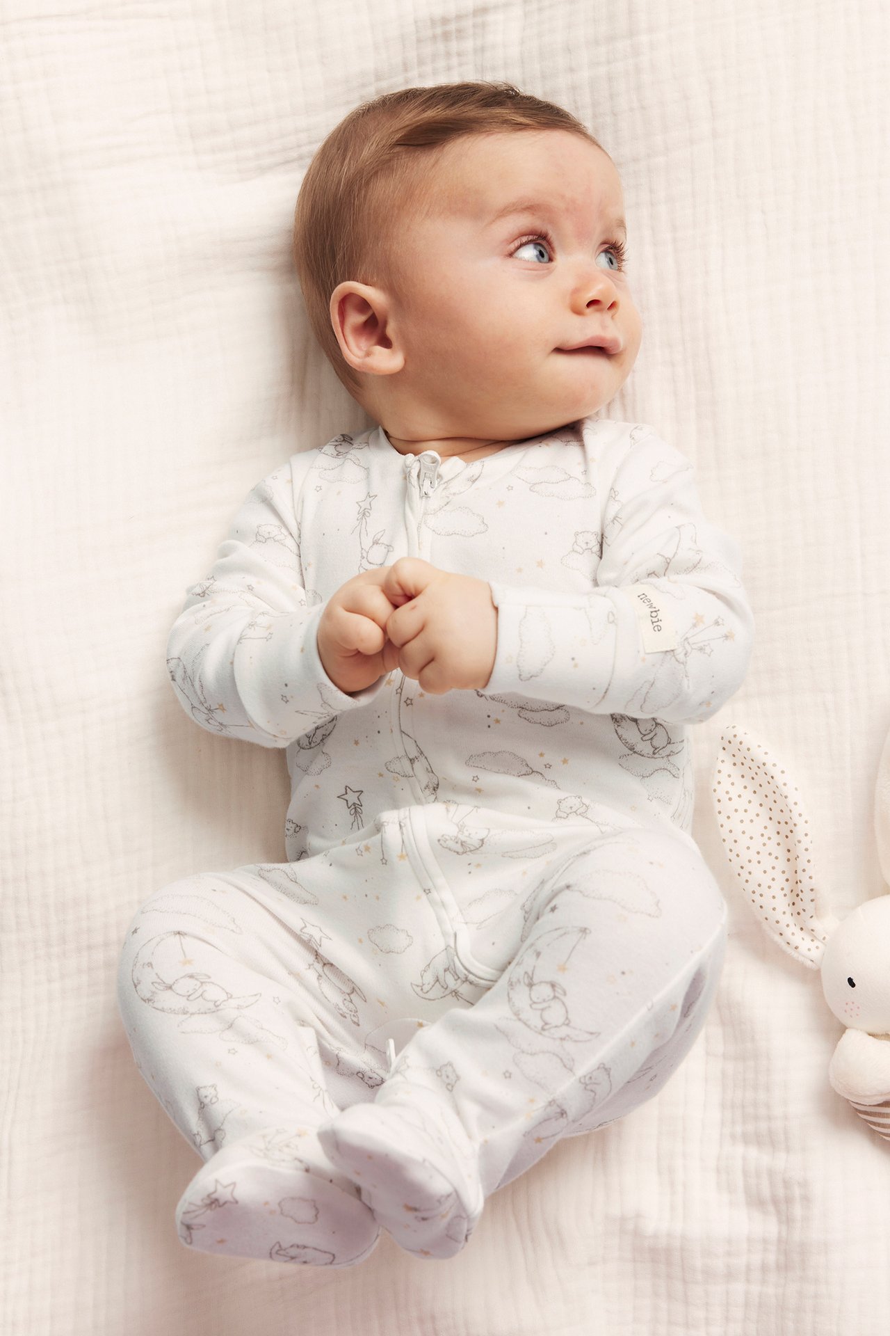 Ylhäältä: vauva offwhite-puuvillapyjamassa, jossa karhu-, kuu-, tähtikuvio, vetoketju, jalkaterät. Kanilelu näkyvissä.