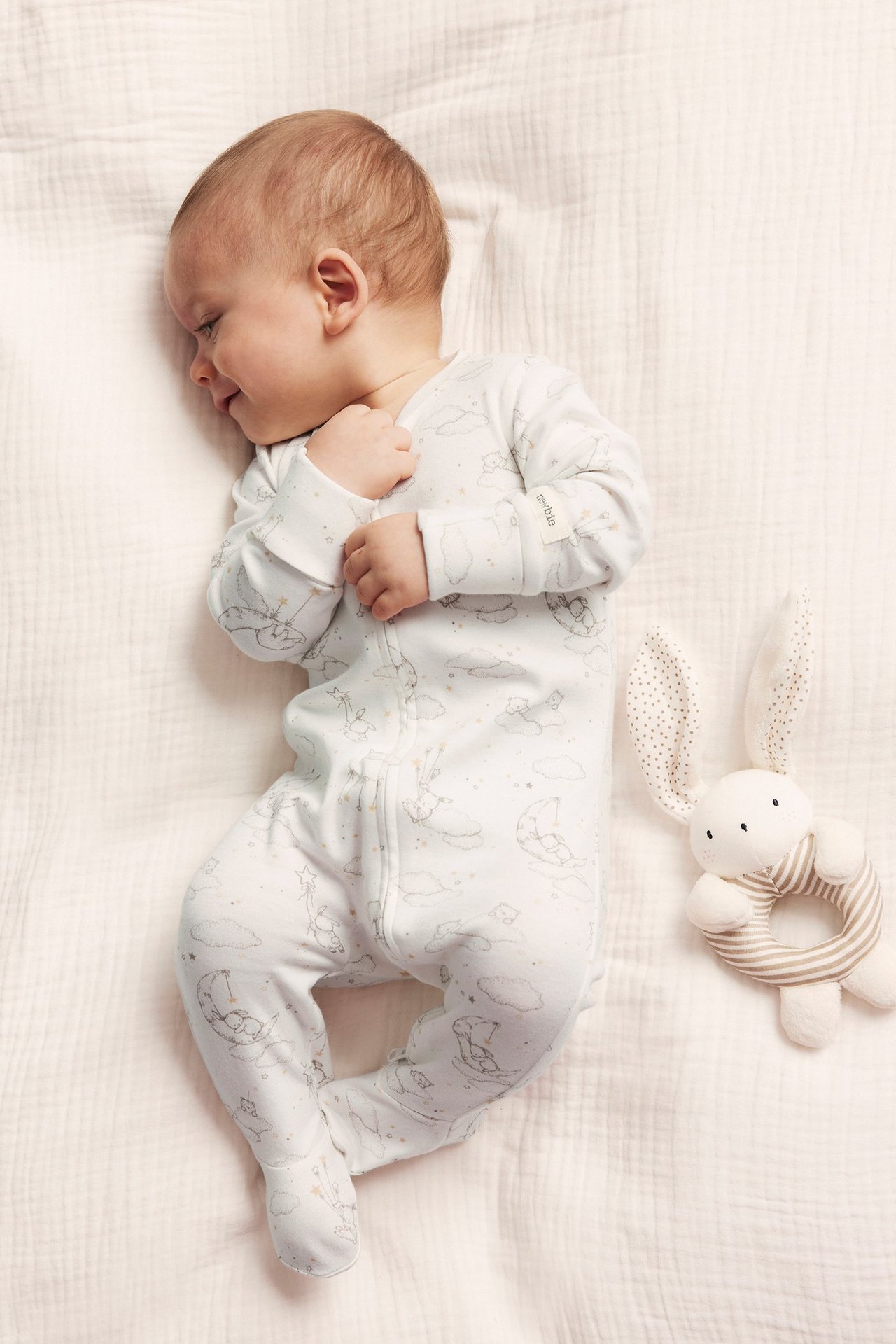 Ovanifrån: baby i offwhite bomullspyjamas med nalle- och månmönster, dragkedja. Kaninskallra bredvid.
