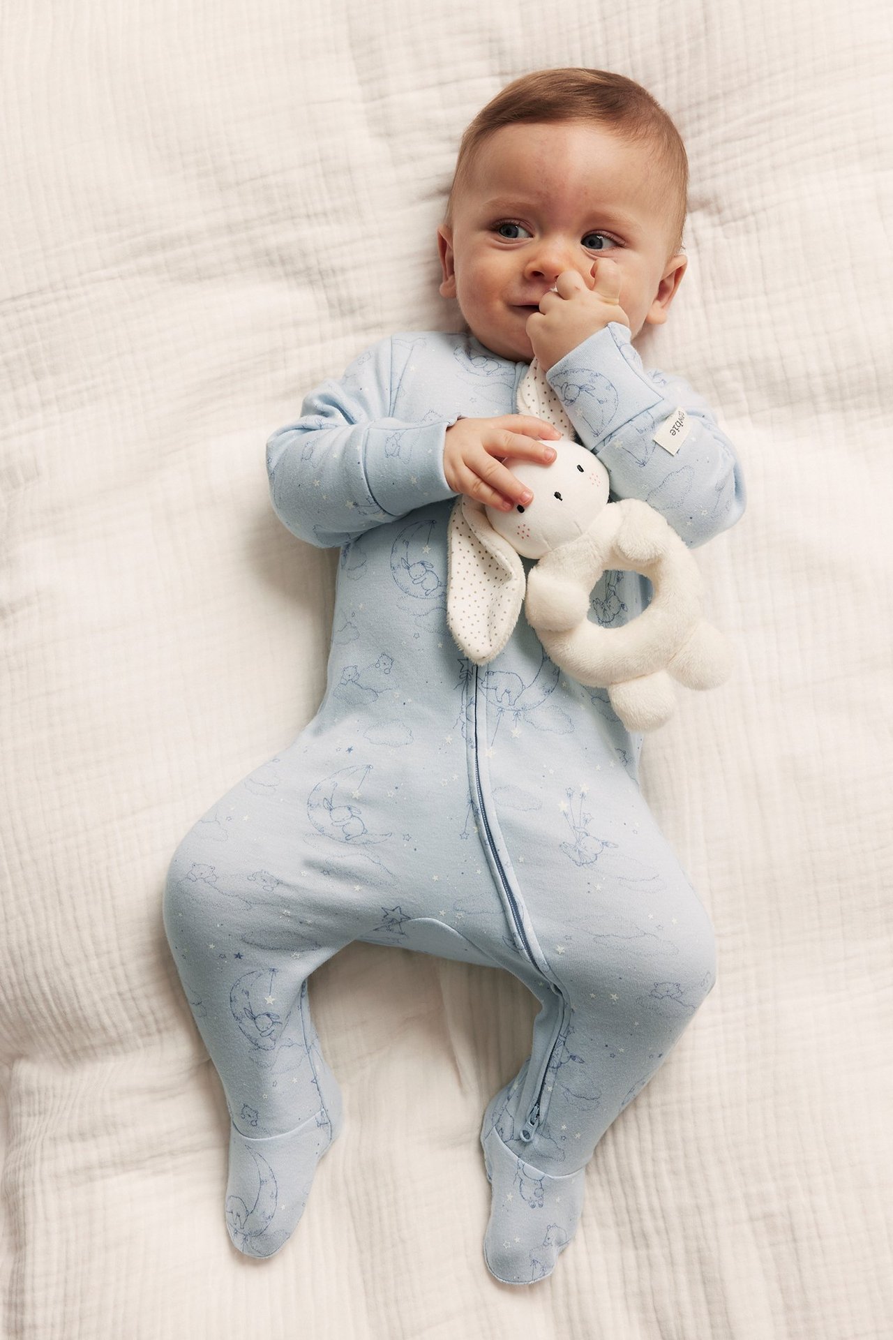 Ylhäältä: vauva yllään vaaleansininen kuviollinen puuvillapyjama vetoketjulla, pitäen valkoista pupuräpätintä.