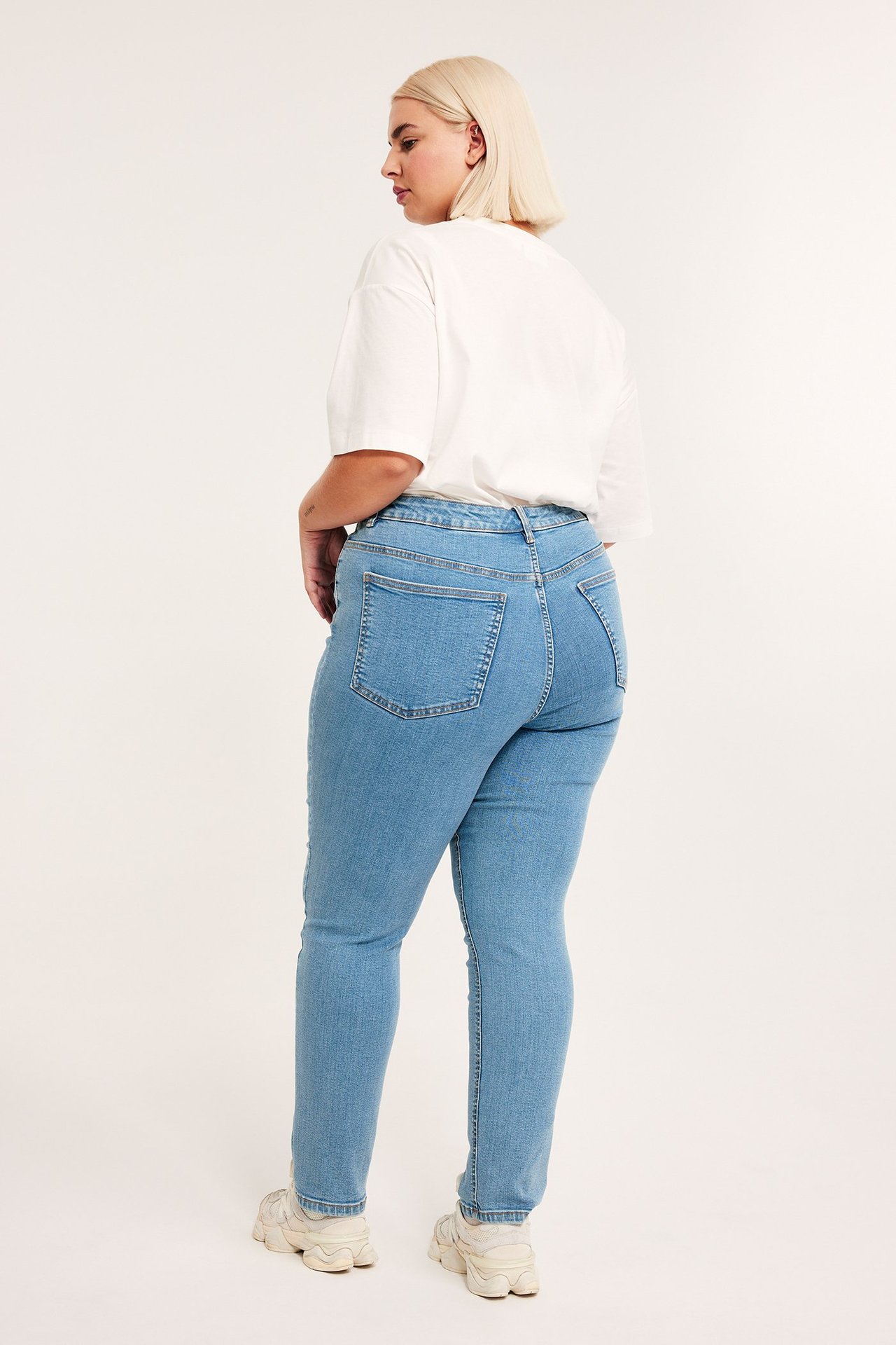 Bakfra: kvinne i lyseblå slim-fit jeans med høyt liv, hvit t-skjorte og beige joggesko.