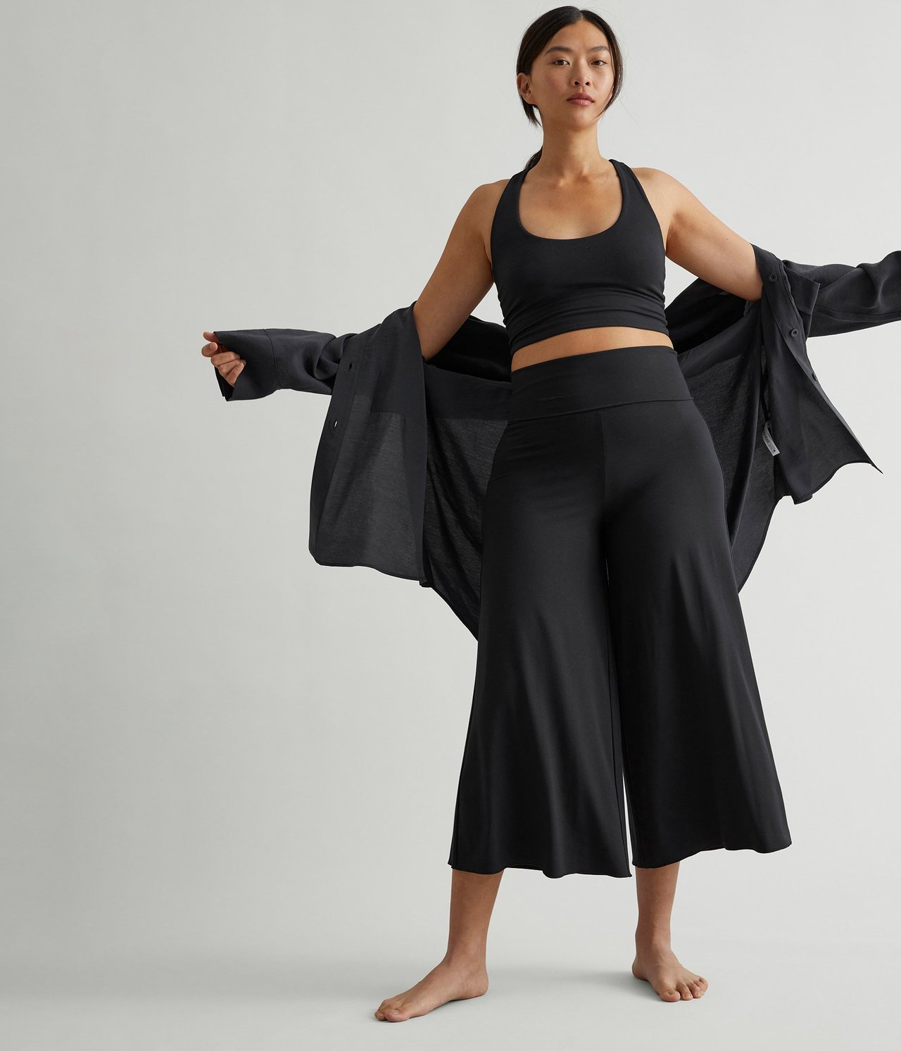 Edestä: nainen mustissa leveälahkeisissa oleskeluhousuissa, mustassa halterneck-topissa ja avoimessa mustassa paidassa.