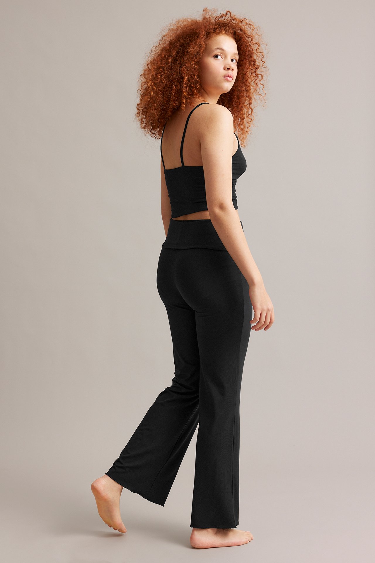 Sidovy: Kvinna i svarta trikå bootcut loungewear-byxor, nedvikbar midja och svart croptop.