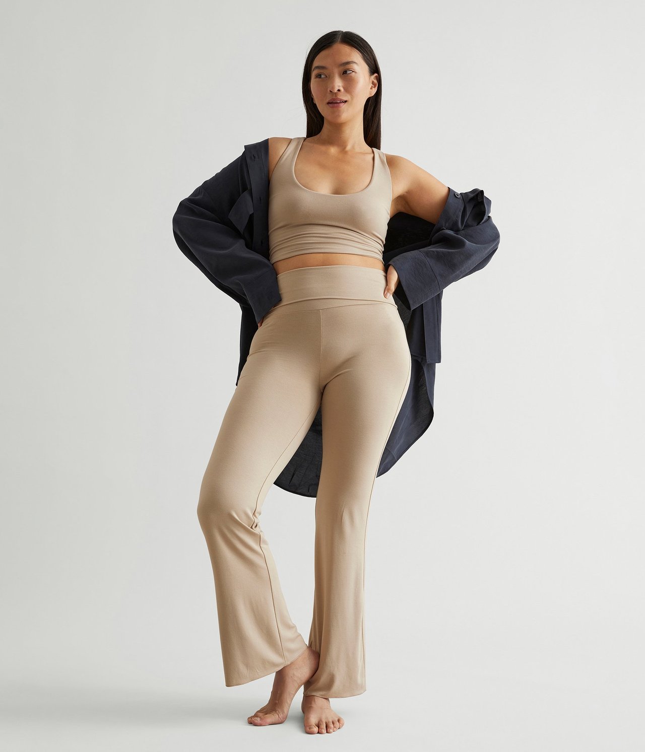 Forfra: Kvinne i lysebrune bootcut-loungewearbukser, matchende crop top og en mørkeblå åpen skjorte.