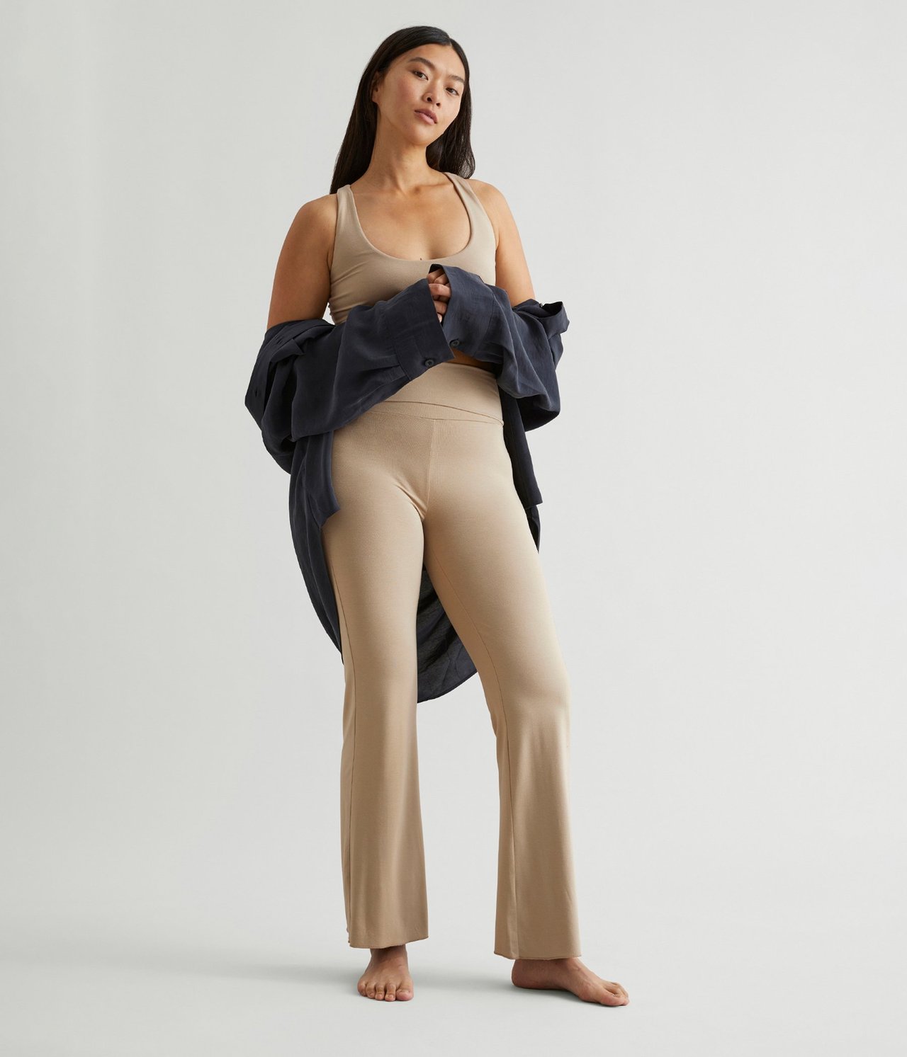 Kvinne som står forfra. Mørkt hår som henger bak skuldrene. Kledd i en beige tanktopp med tynne skulderstropper. Beige utsvingte leggings som matcher toppen. Mørkeblå skjorte som henger over armene.