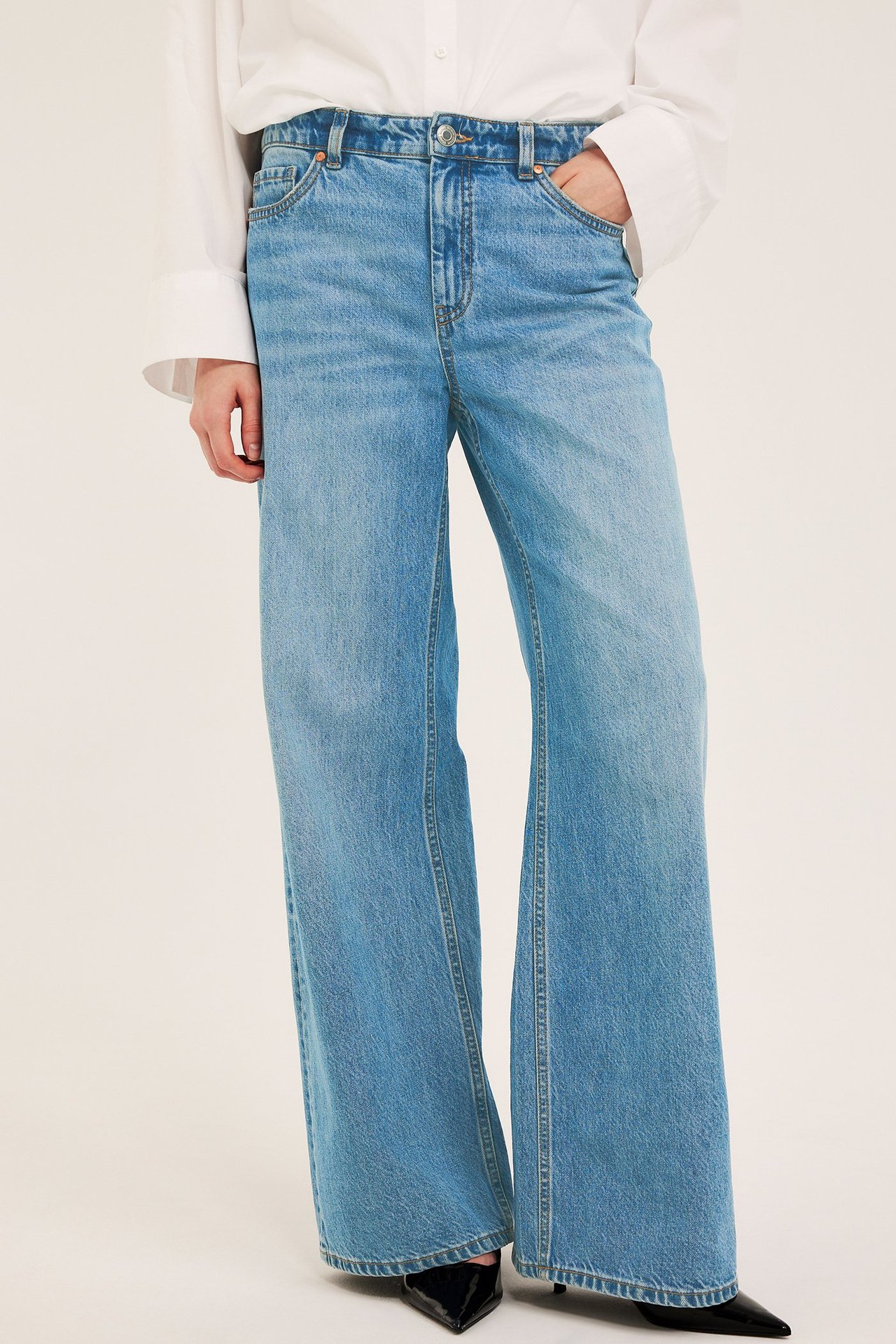 Framsida av kvinna i ljusblå höga vida jeans, vit skjorta och svarta spetsiga klackskor.