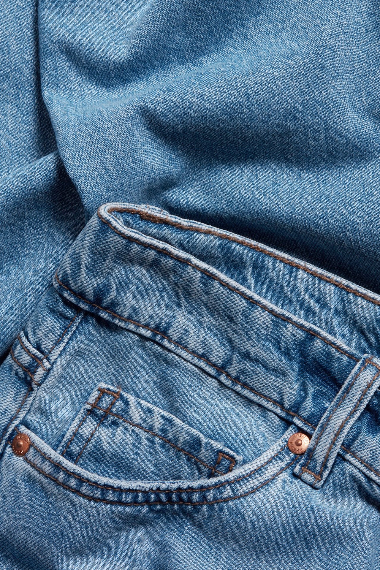 Zbliżenie jasnoniebieskich jeansów z sztywnego denimu, widoczny pas, szlufki, przednia kieszeń i miedziane nity.