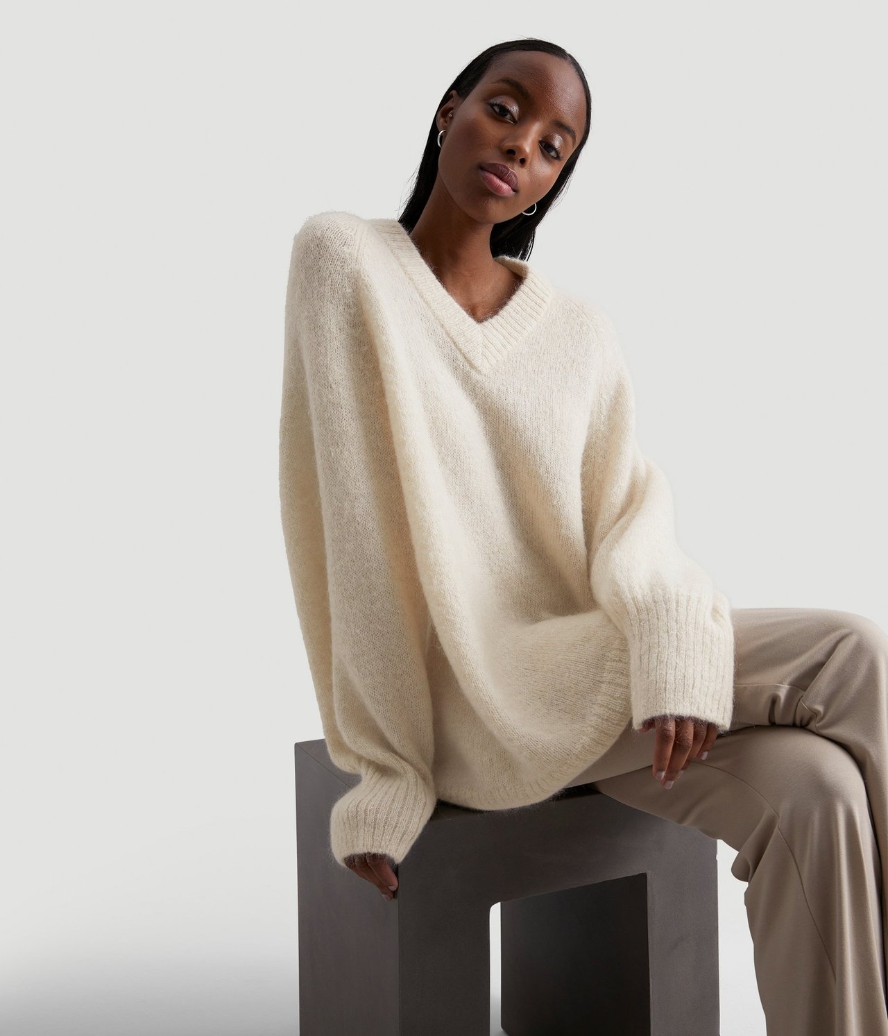 Forfra: Kvinne i en oversized lys beige melert V-hals ullmiks genser og beige bukser.