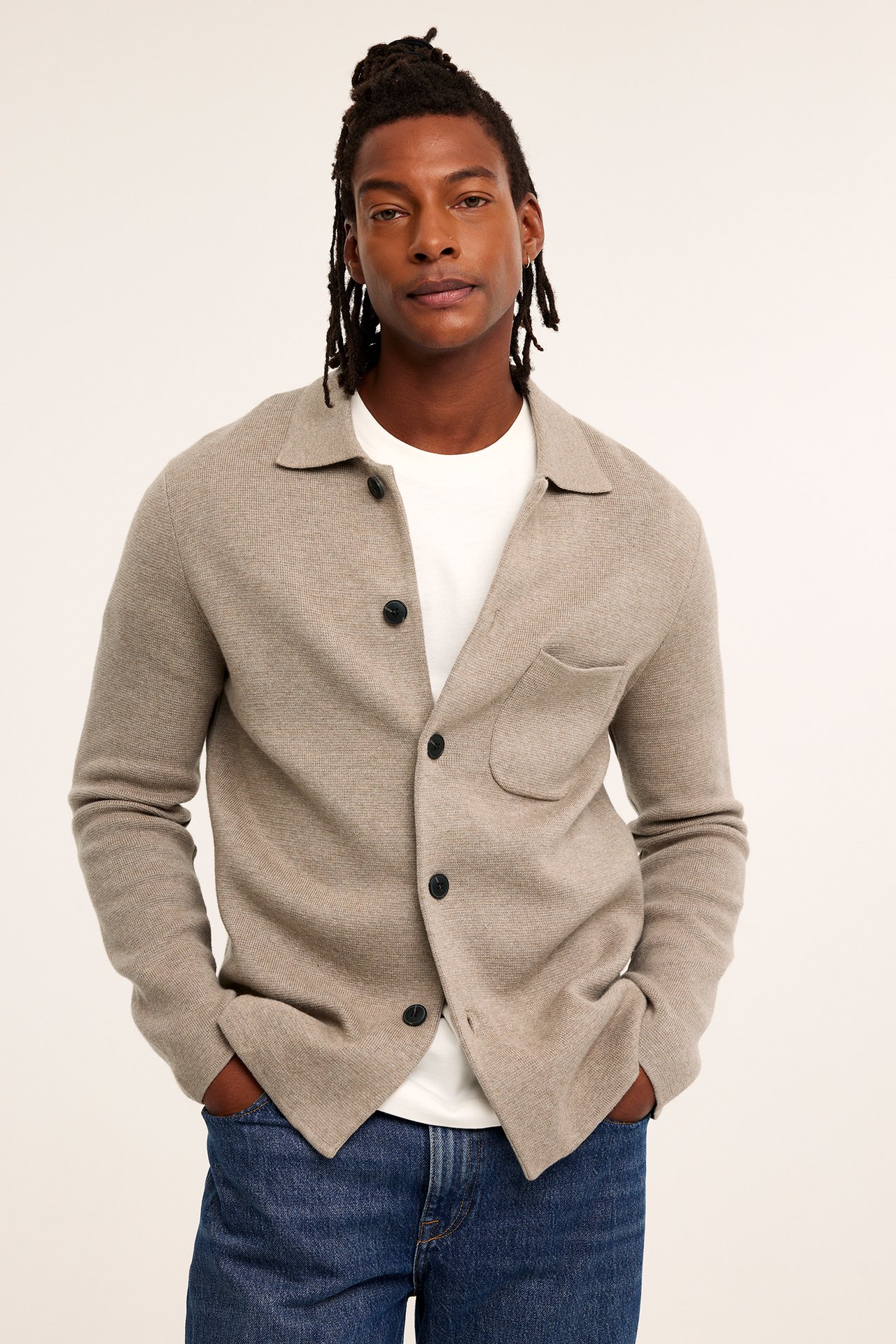 Mann i beige melert strikket cardigan med krage, brystlomme, knapper, hvit t-skjorte, blå jeans.