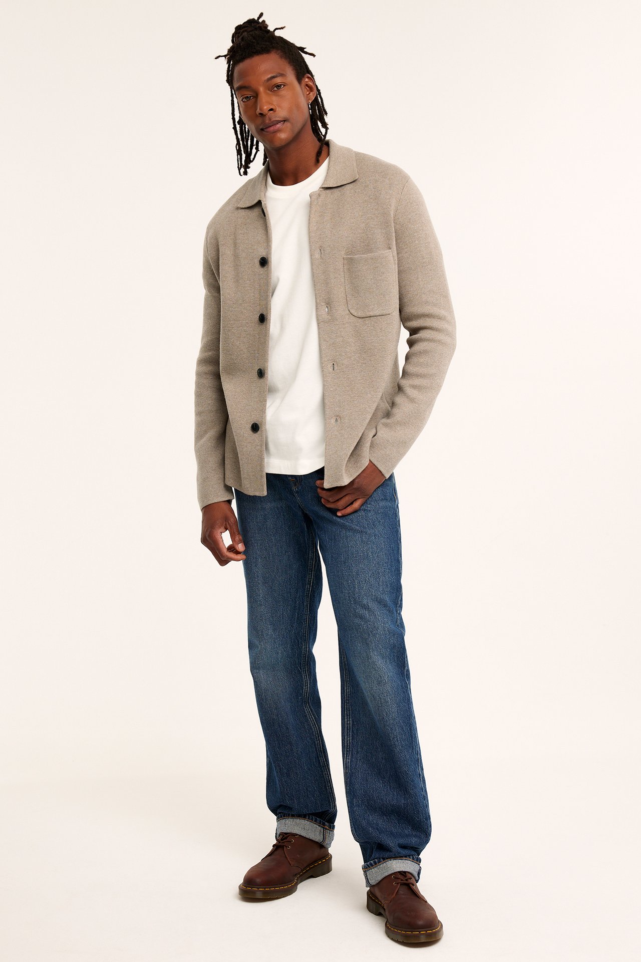 Forfra: Mann i beige melert strikket cardigan med krage, brystlomme, hvit t-skjorte, jeans, brune sko.