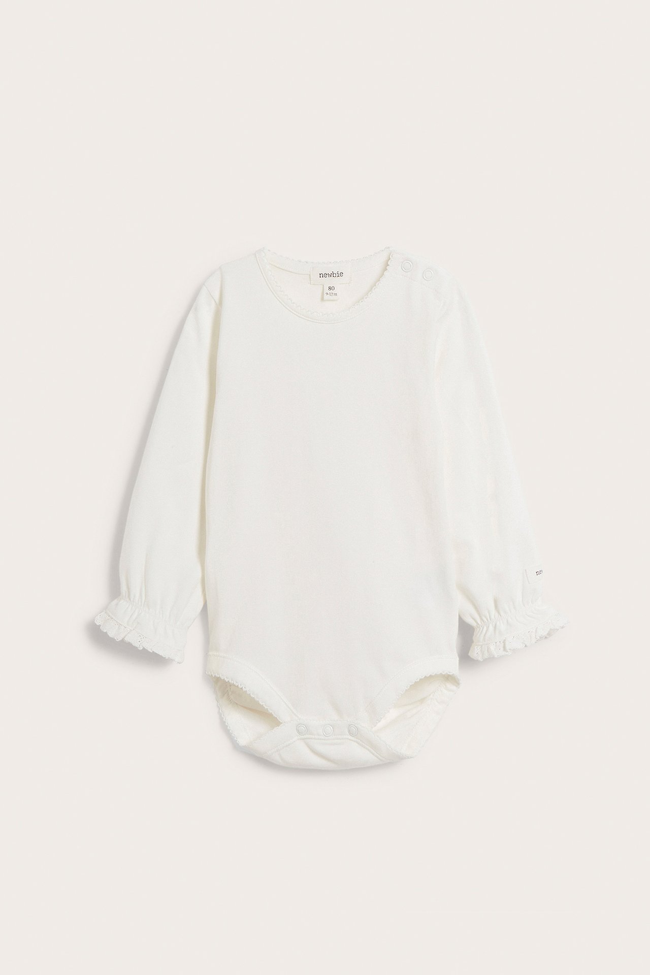 Frontbilde av offwhite langermet babybody med picot-kant i halsen, blonde-volang mansjetter og trykknapper.