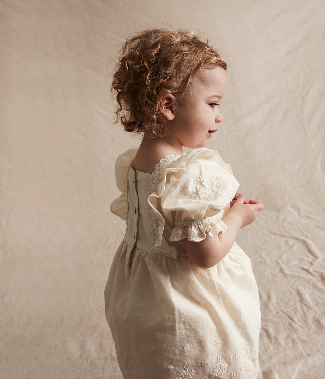 Sidovy av en baby i offwhite broderad bomullsklänning med puffärmar och knappar bak.