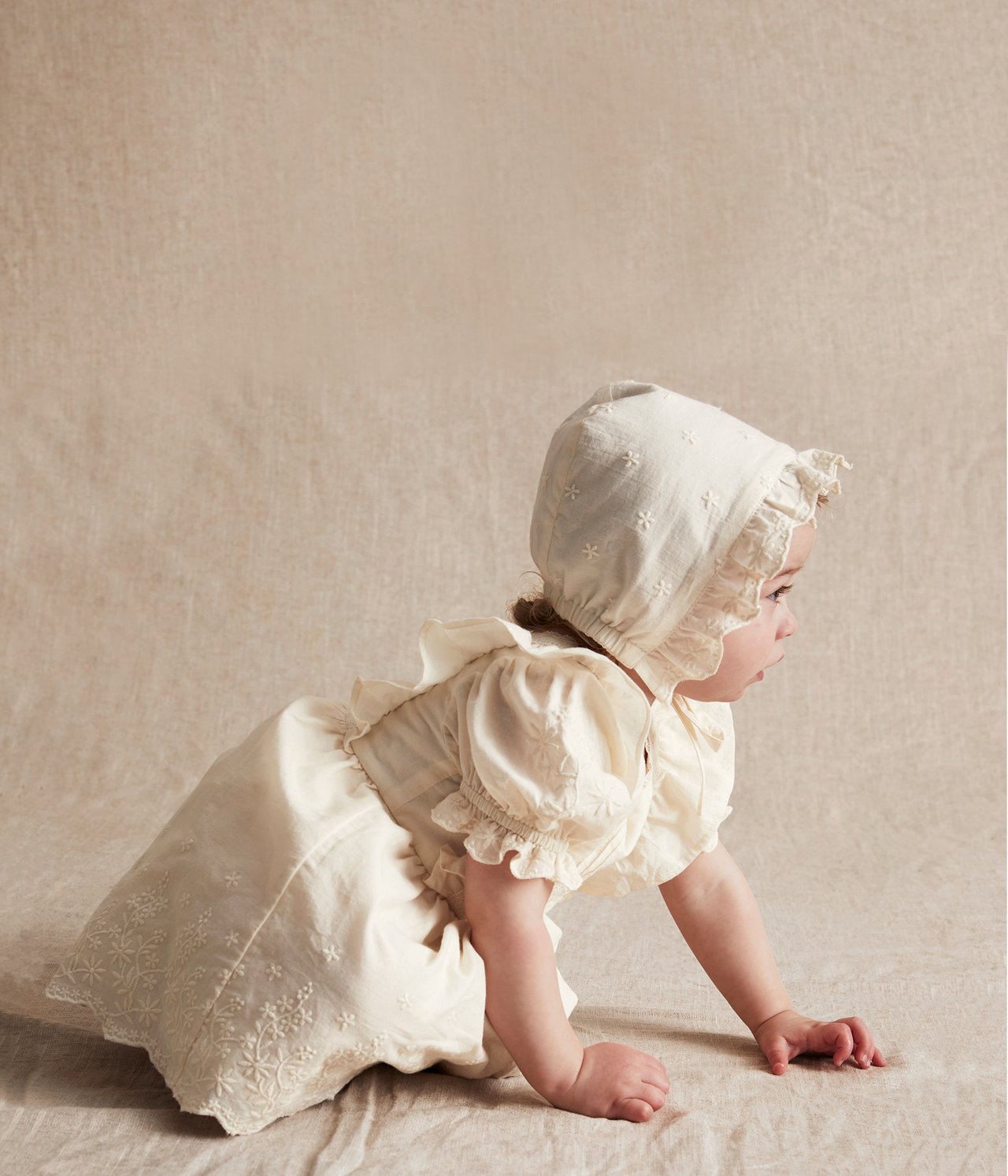 Sidovy av en baby som kryper i en offwhite broderad bomullsklänning med puffärm och matchande mössa.