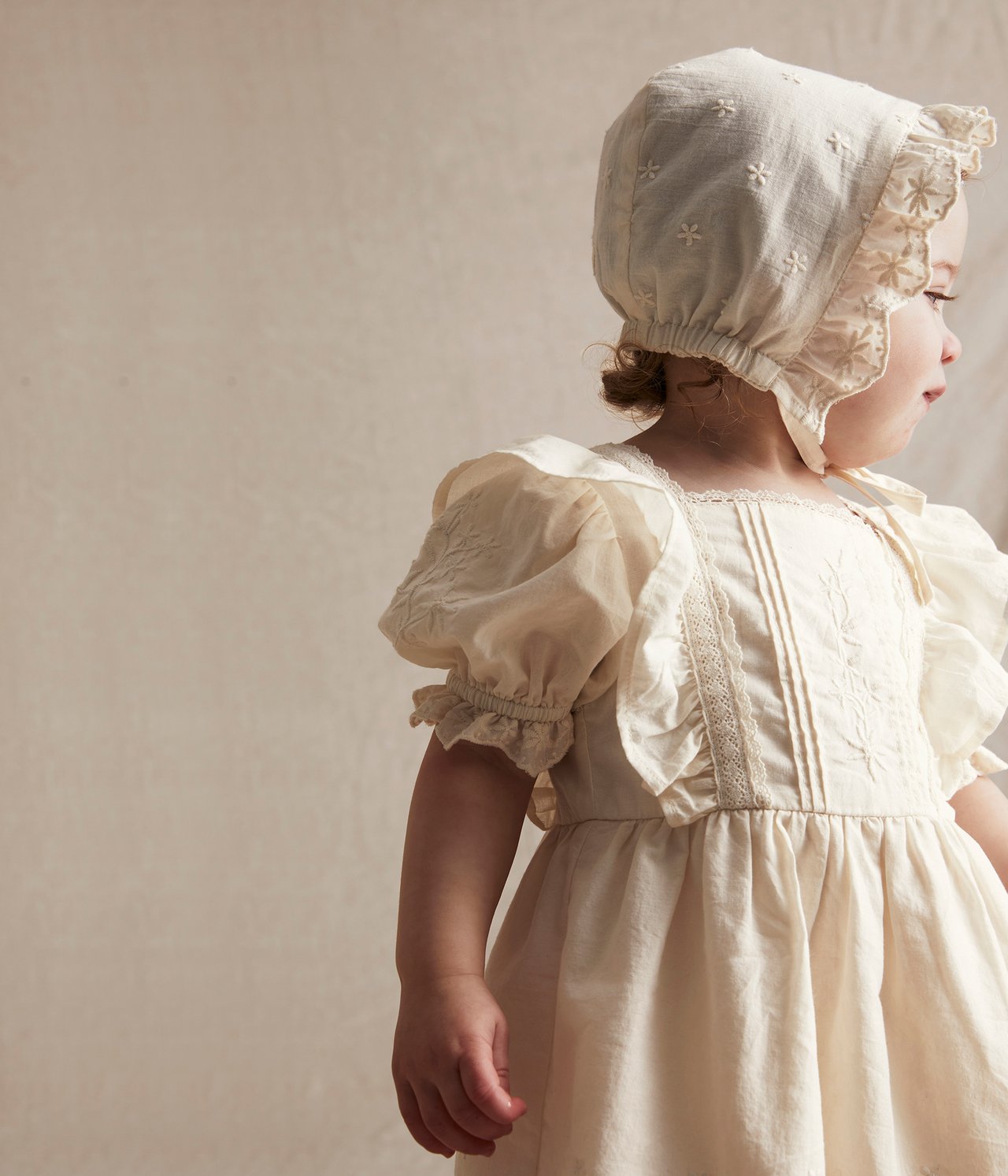 Sidovy av en baby i en naturvit broderad bomullsklänning med puffärmar och matchande mössa.
