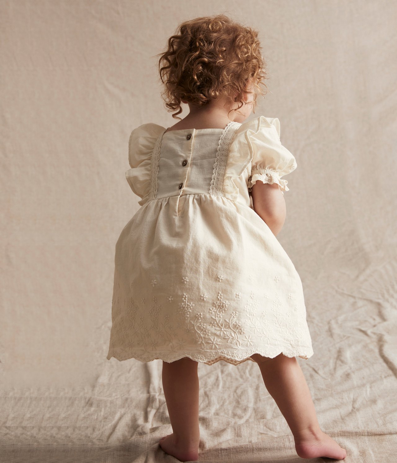 Bakifrån, baby i offwhite broderad bomullsklänning med puffärmar, spetsdetaljer och knappar bak.