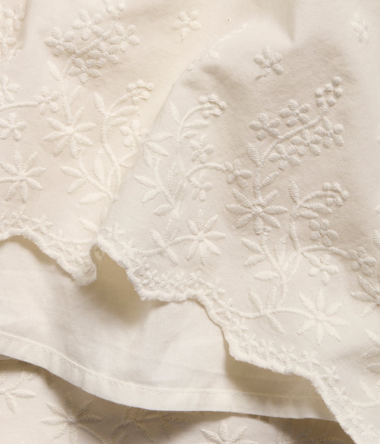 Närbild av offwhite vävd bomull med intrikata blombroderier och vågig spetskant.