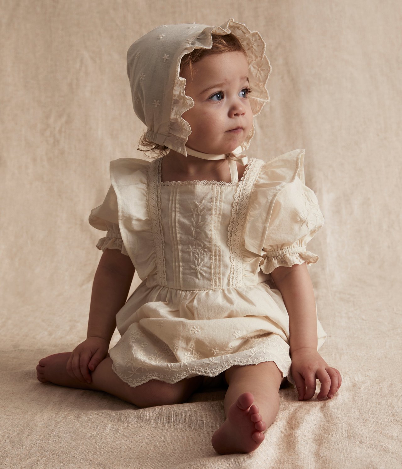 Framifrån: baby i offwhite broderad bomullsklänning med puffärm och matchande hätta.