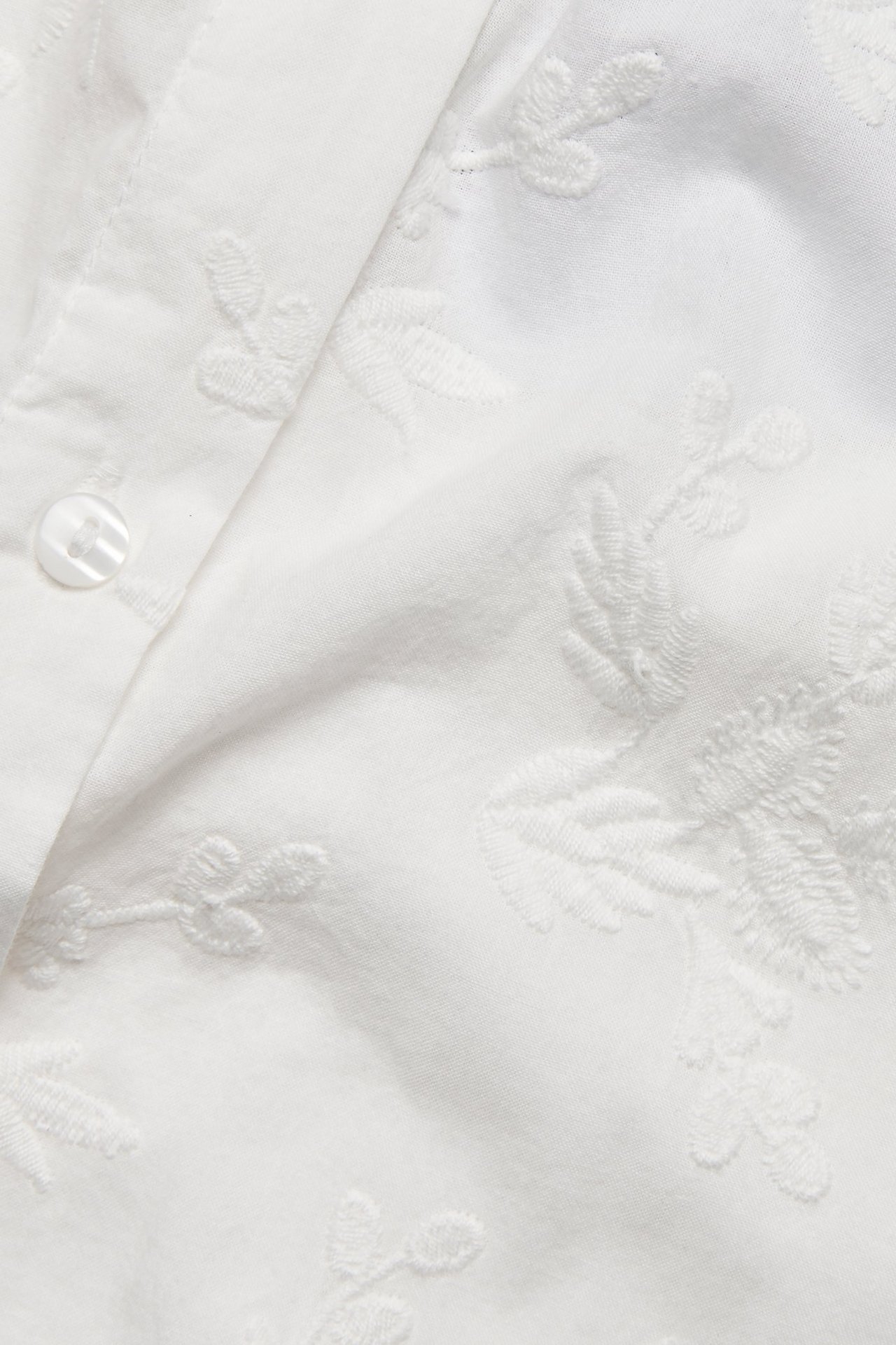 Zbliżenie na ecru bawełnianą bluzkę z haftowanymi wzorami kwiatowymi i widocznym guzikiem.