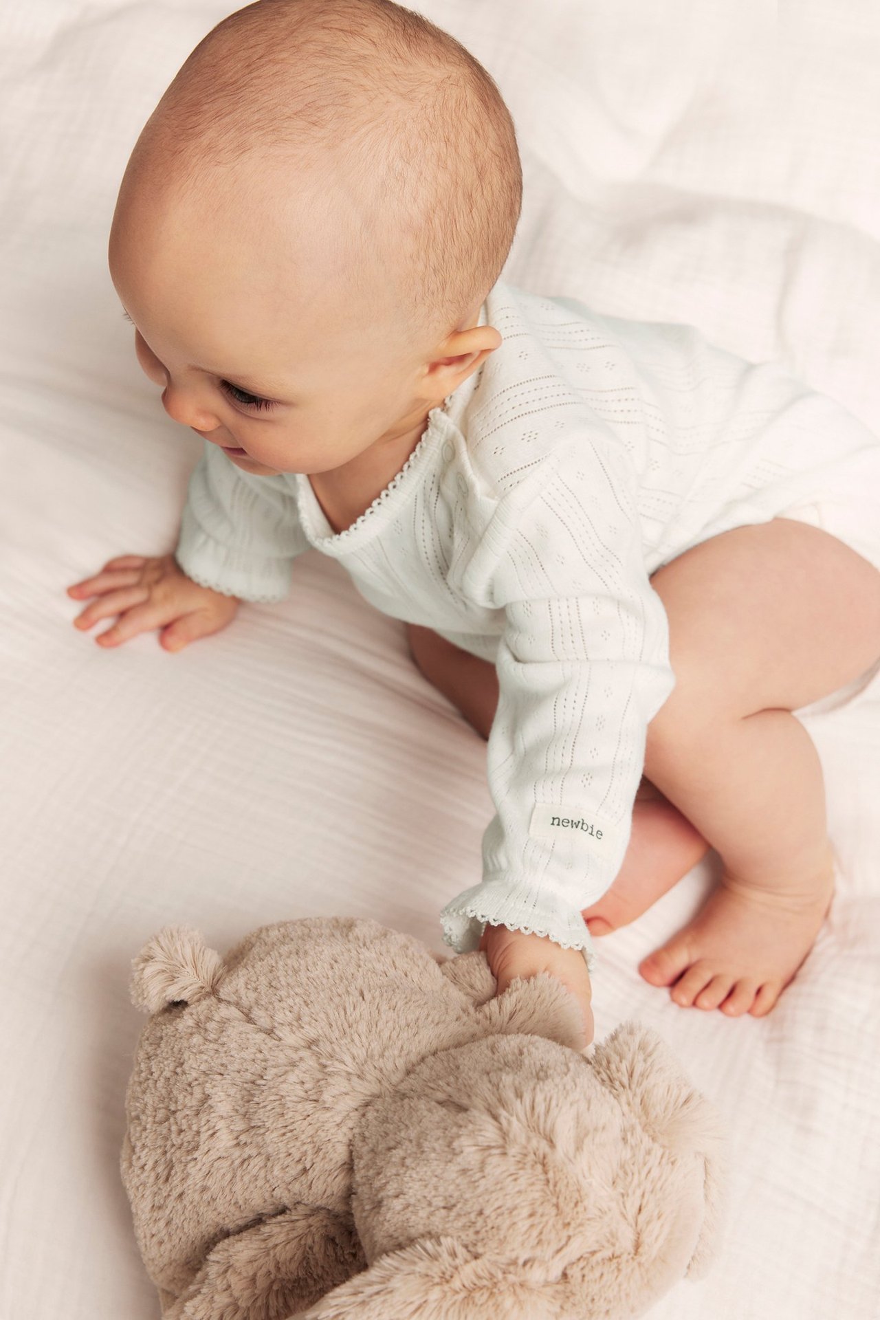 Ylhäältä näkymä vauvasta luonnonvalkoisessa pointelle-ribatussa bodyssä, koskettaen beigeä nallekarhua.