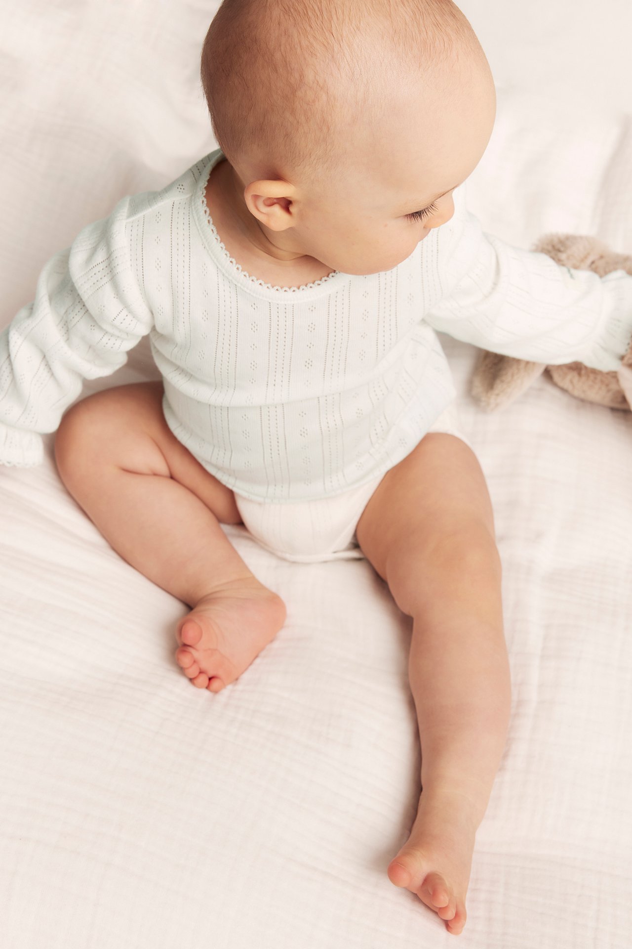 Yläkuva vauvasta yllään luonnonvalkoinen pointelle-ribattu body picot-reunuksella, pitäen pehmolelua.