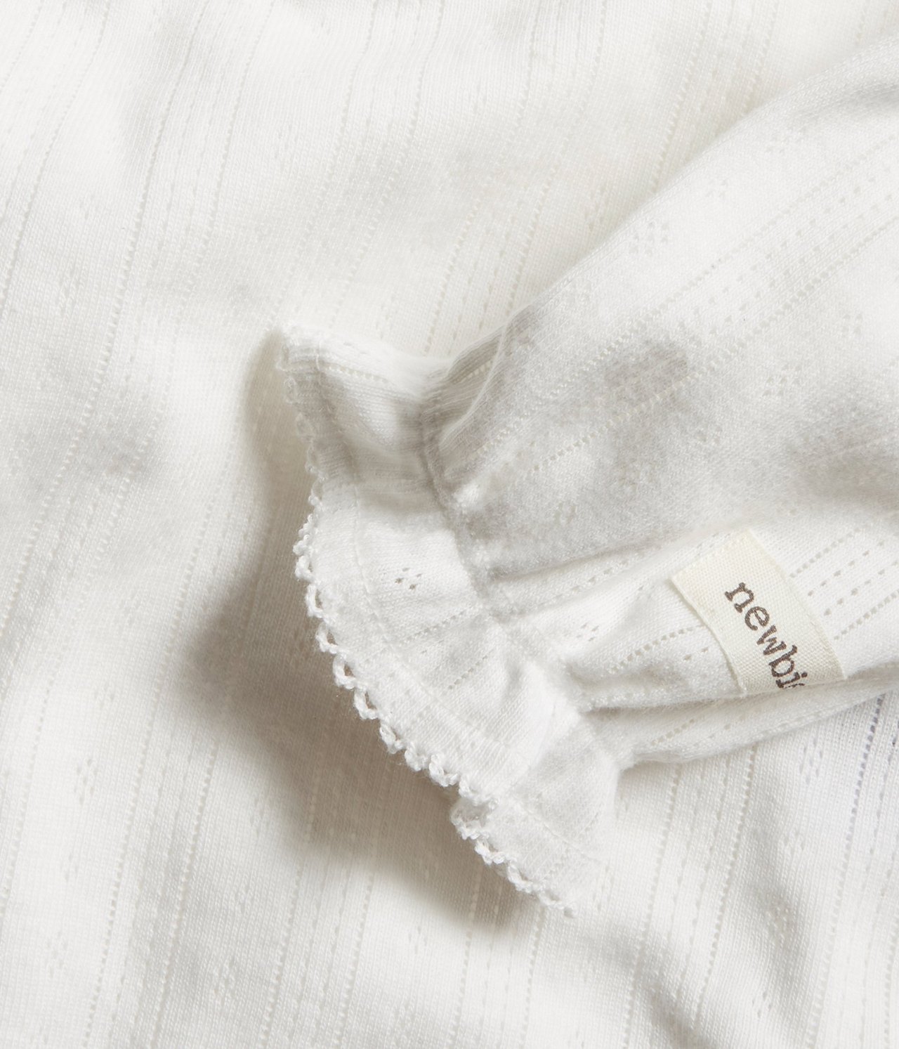 Zbliżenie na ecru body niemowlęce z organicznej bawełny w prążki pointelle, z falbanką i wykończeniem picot.