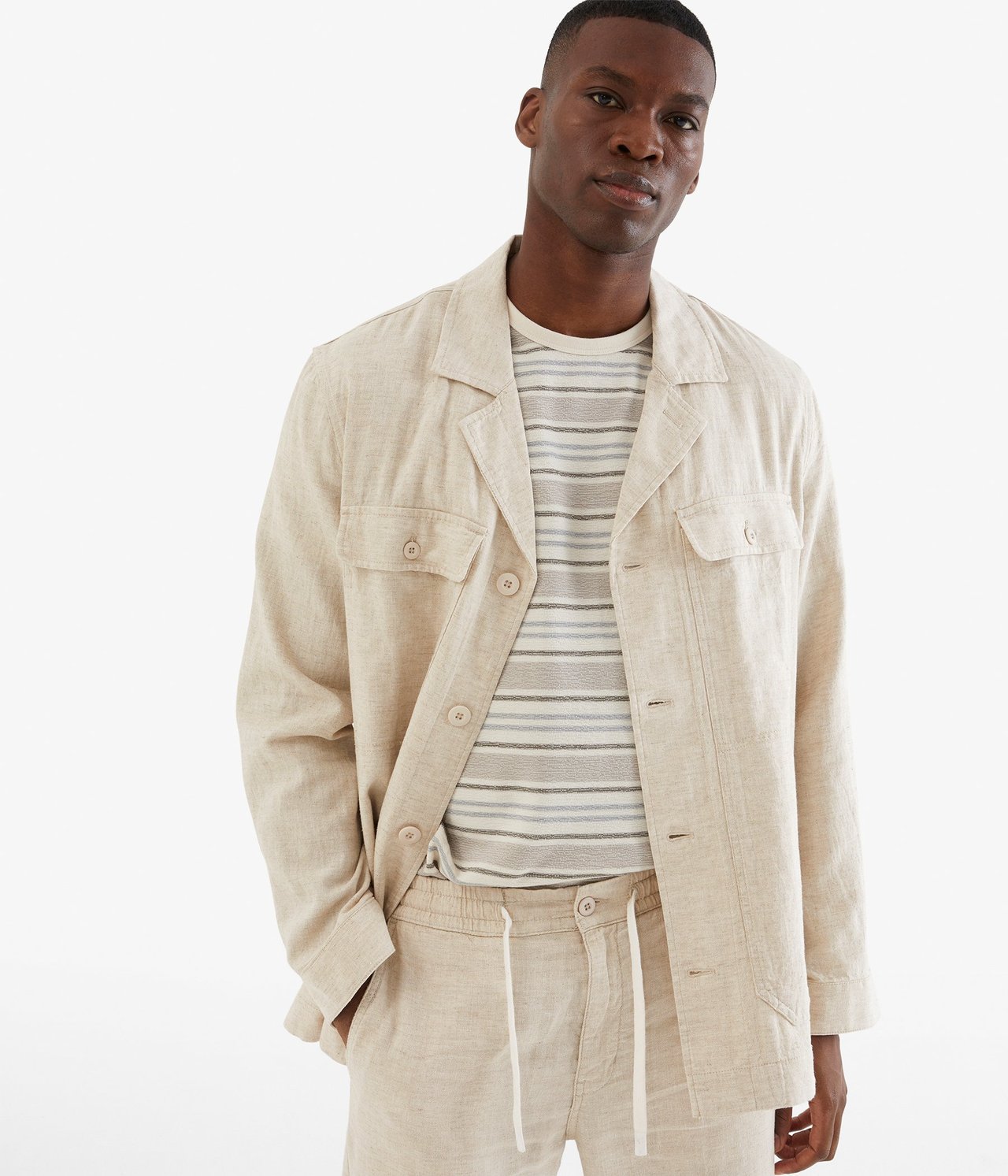 Widok z przodu mężczyzny w rozpiętej beżowej koszuli-kurtce z lnu, pasiastym t-shircie i pasujących beżowych spodniach.