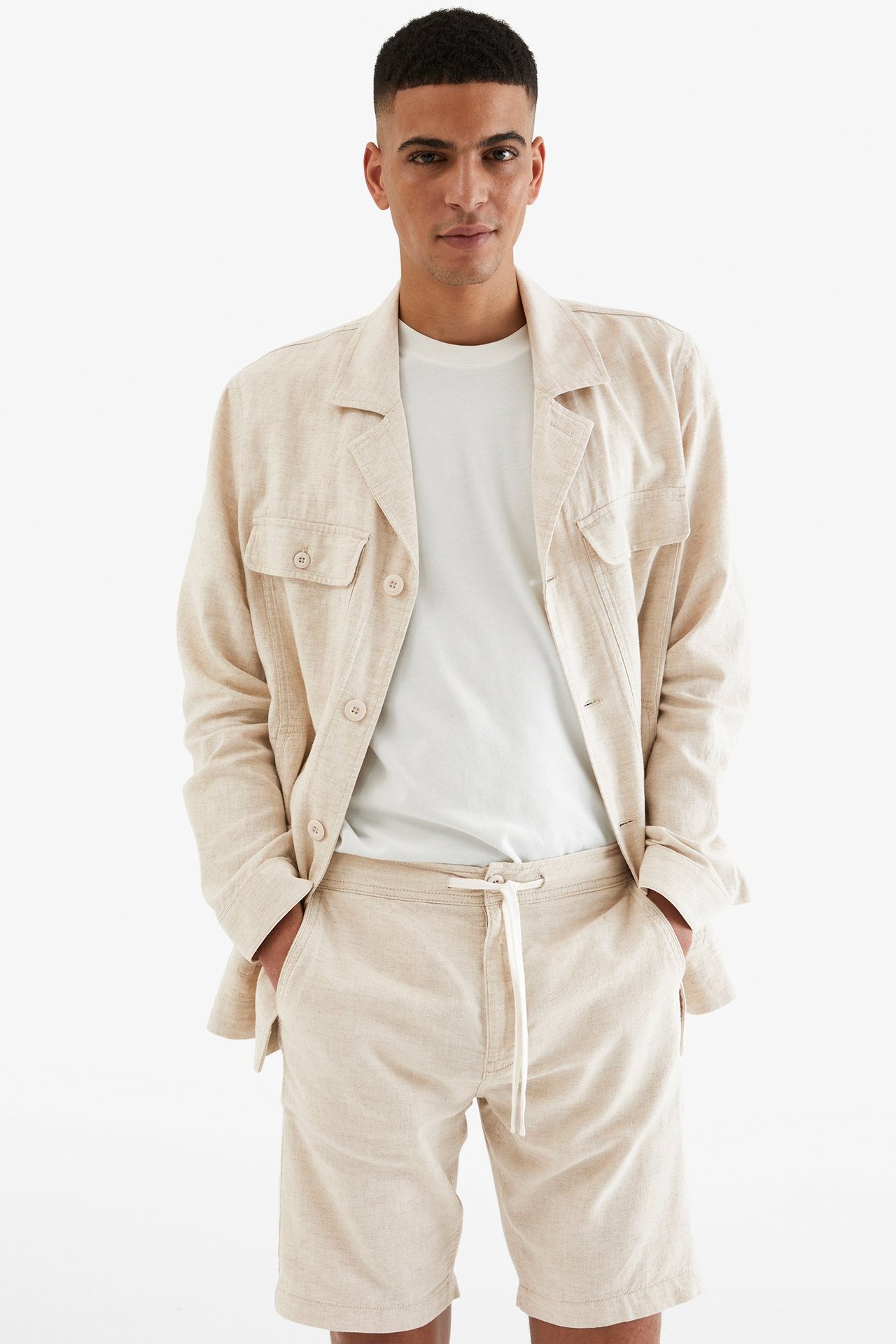 Edestä: Mies vaaleanbeiget pellavasekoiteshortsit, kiristysnyöri, valkoinen t-paita ja avoin vaaleanbeige pellavatakki.