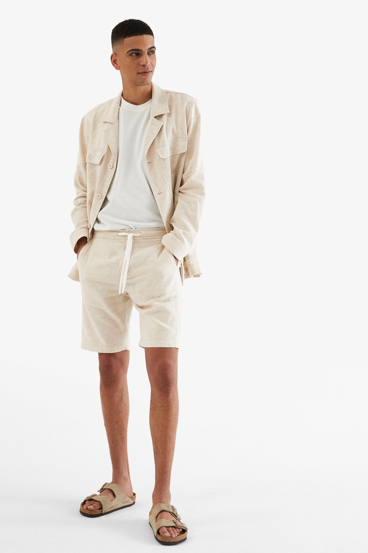 Mies yllään vaaleanbeiget pellavasekoiteshortsit kiristysnyörillä, valkoinen t-paita, yhteensopiva paitatakki ja beiget sandaalit.